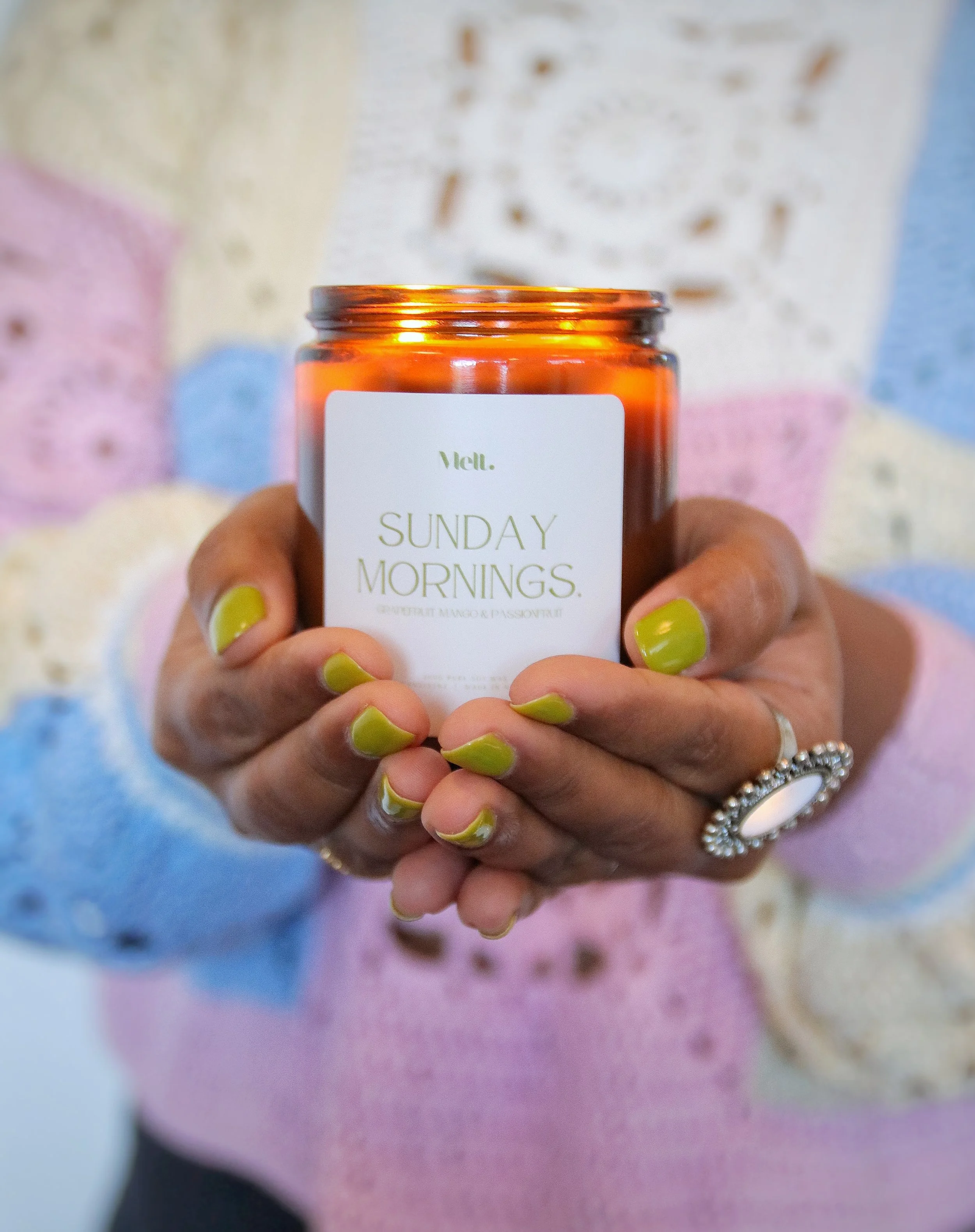 A person with yellow-green painted nails holding a jar labeled 'Sunday Mornings' scented candle, with a colorful background.
