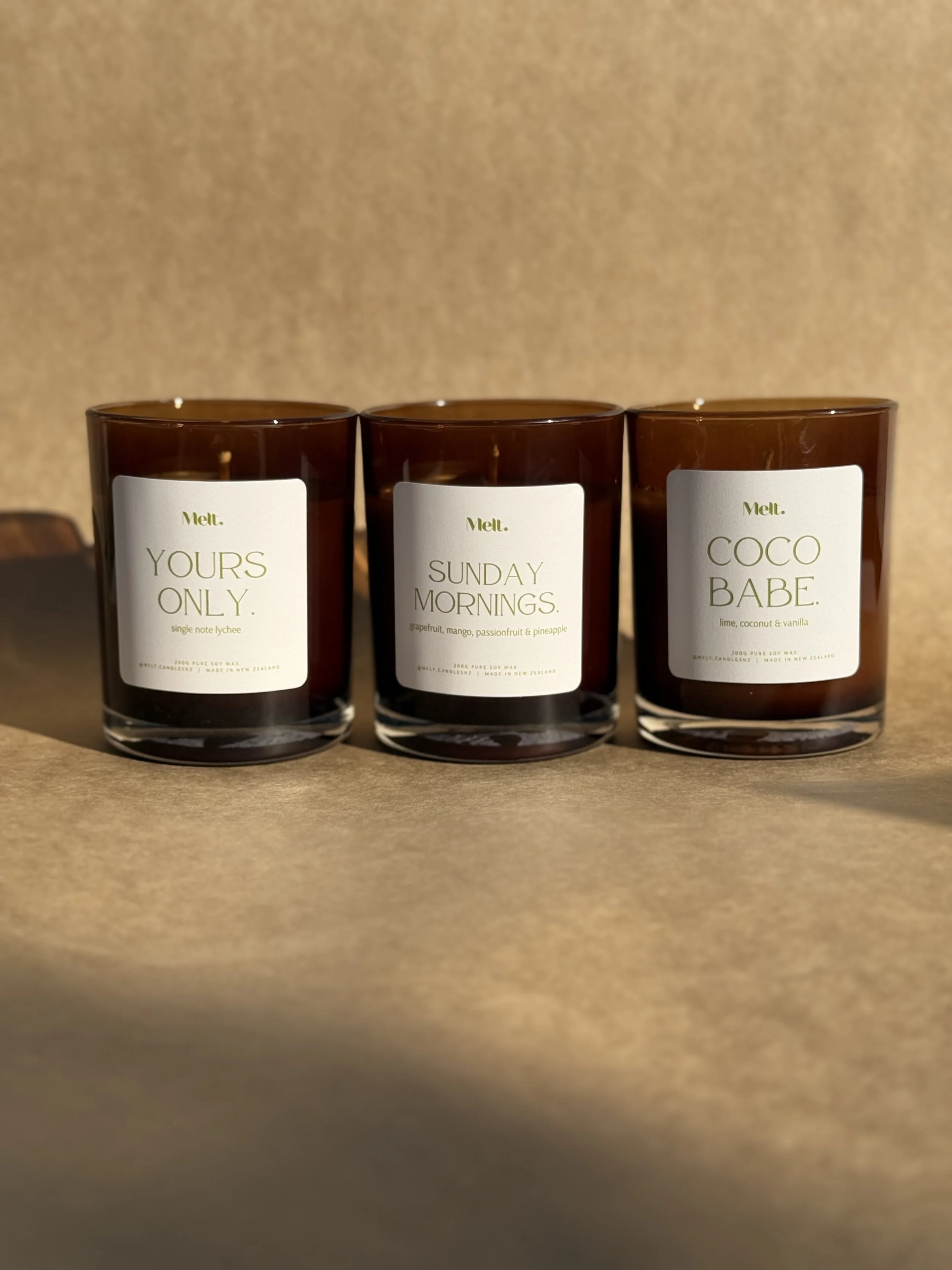 Three scented candles in dark brown glass containers with white labels, each with different names and scents. From left to right: 'Yours Only,' 'Sunday Mornings,' and 'Coco Babe.'