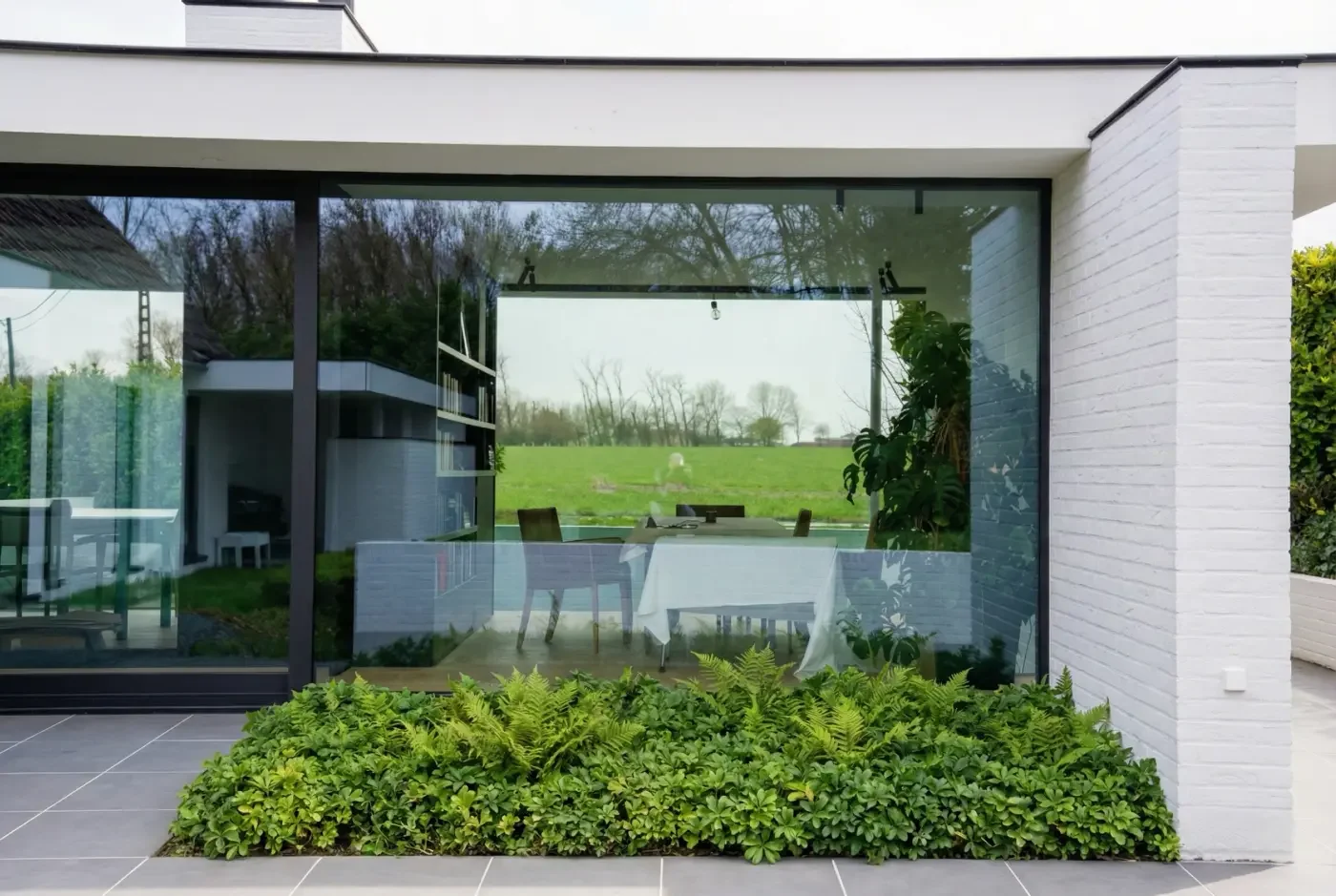 View of a modern house with large glass windows, a white brick wall, and a garden with green bushes in the foreground. Through the window, an outdoor dining table and chairs are visible, with a grassy field and trees in the background.