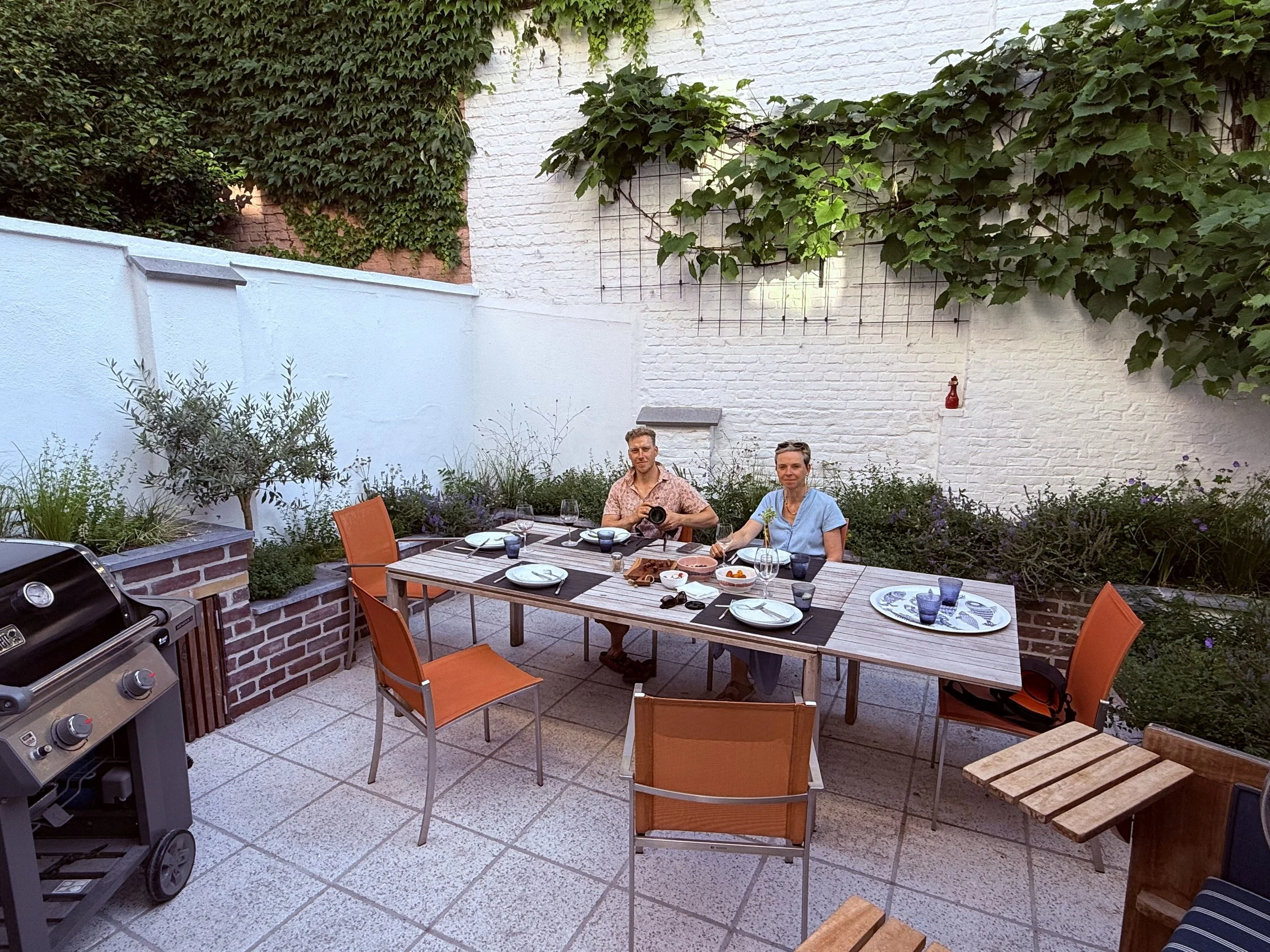Een sfeervol ingerichte stadstuin met een grote houten eettafel, oranje stoelen, een barbecue en witgekalkte muren begroeid met klimplanten.