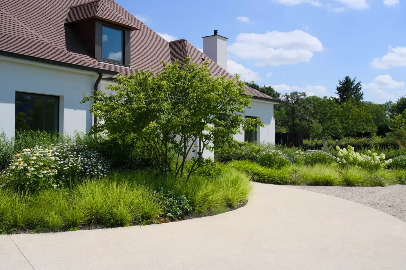 A modern house with a reddish-brown roof, white walls, and large windows, surrounded by lush greenery and flowering plants under a partly cloudy sky.
