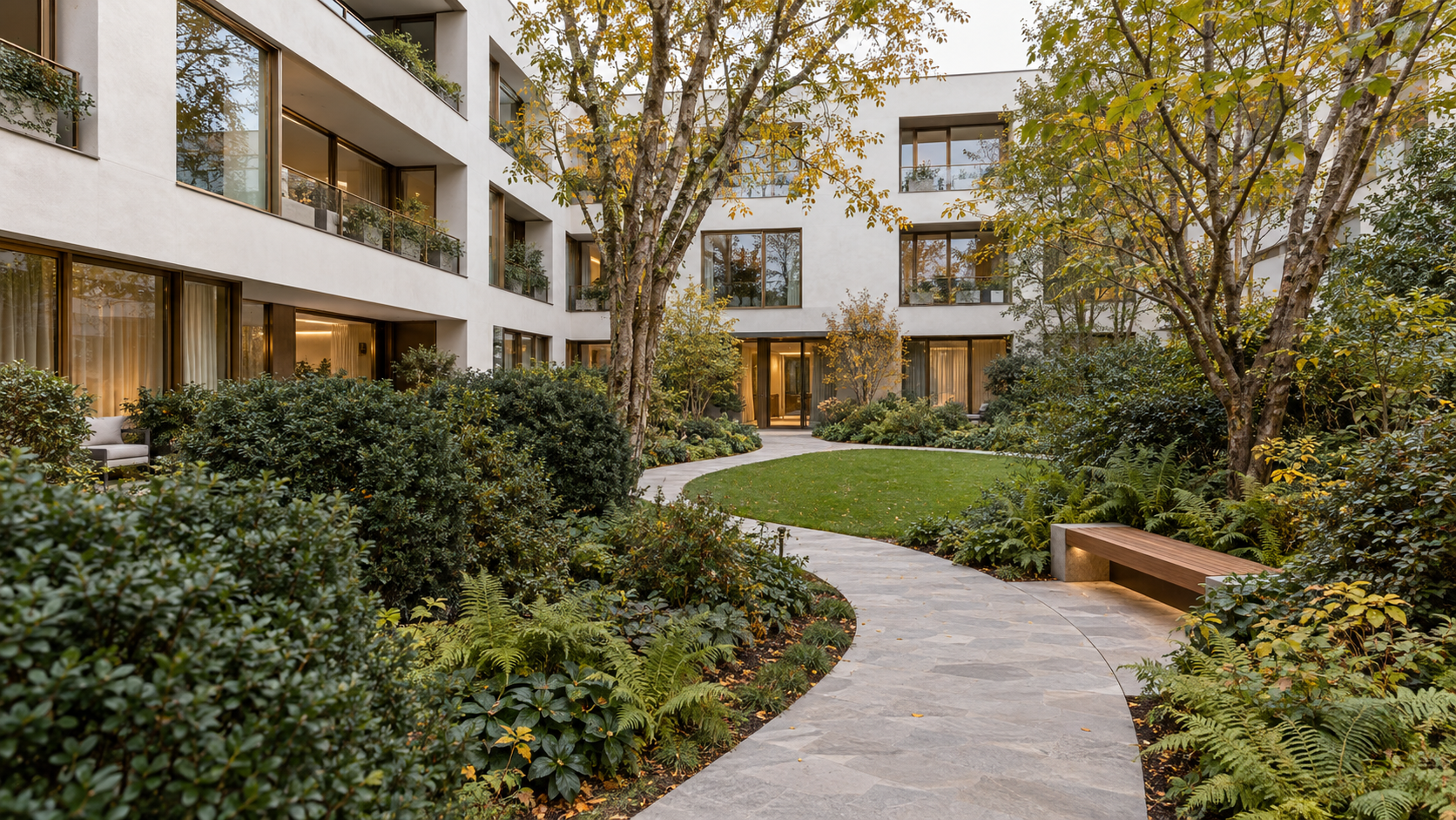 Gemeenschappelijke tuin met volwassen bomen, groene struikgroepen en een vrij wandelpad tussen appartementsgebouwen.
