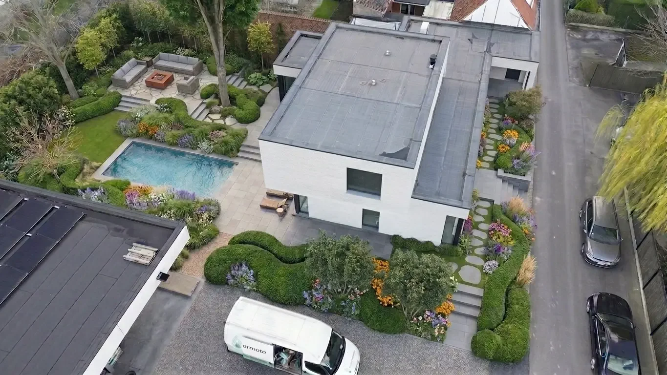 Aerial view of a modern house with a landscaped backyard, swimming pool, sitting area, and variety of plants and flowers surrounding the property.