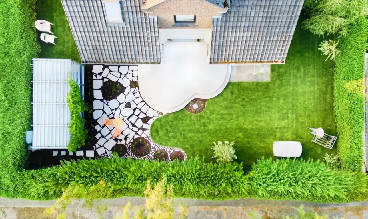 Overhead view of a backyard with a grassy lawn, a paved patio, a white landscaped area with irregular stones, a shed, a white cylindrical object, and a barbecue grill, all surrounded by tall green hedges and trees.