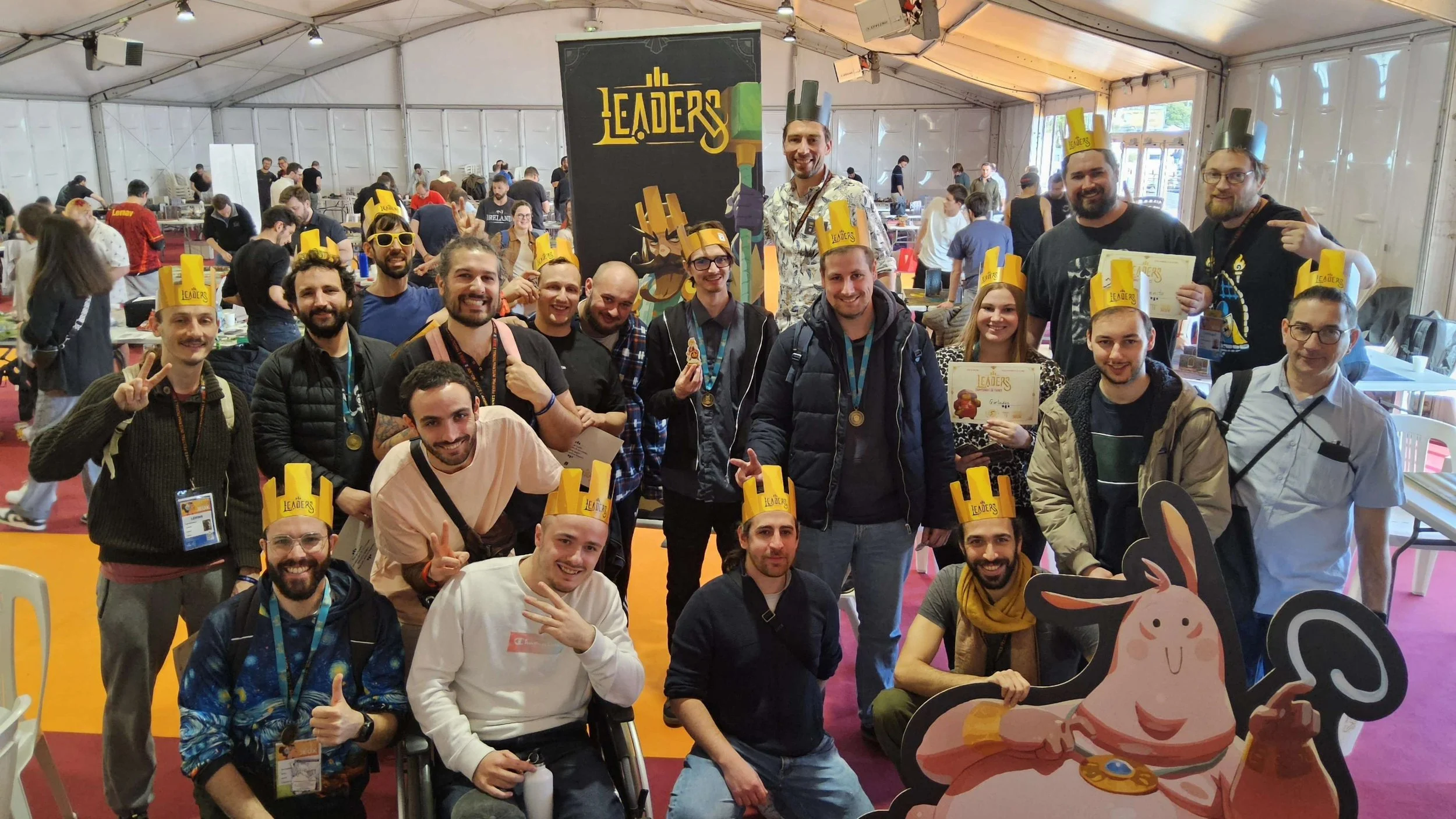 Groupe de personnes lors d'un événement, portant des chapeaux en carton portant l'inscription 'LEADERS', posant pour une photo avec des sourires et faisant des gestes de paix. La scène se déroule dans une grande tente, avec beaucoup d'autres participants en arrière-plan.