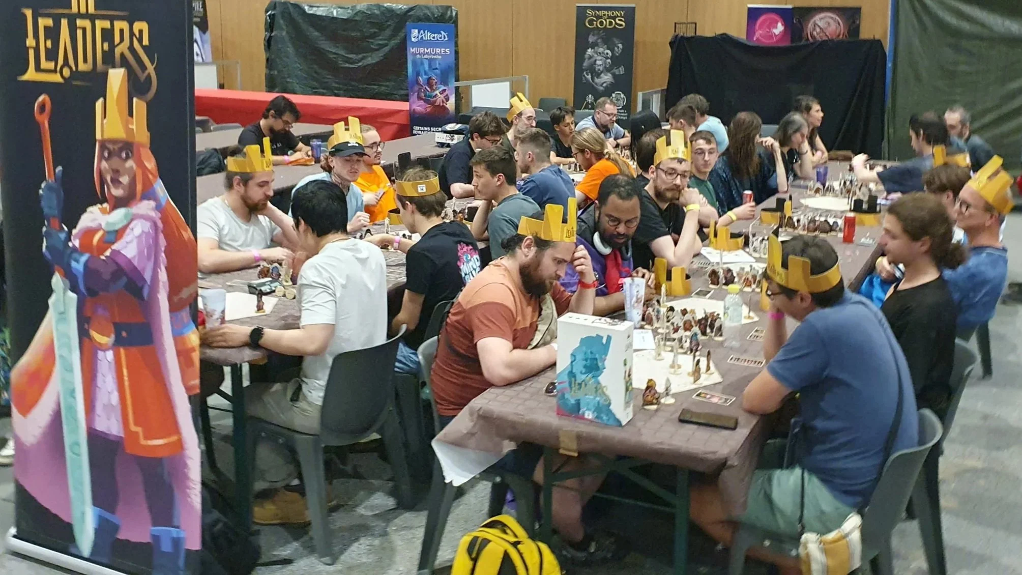 Une salle remplie de personnes jouant à un jeu de société, portant des couronnes en papier. Le décor évoque une ambiance de tournoi ou de compétition avec des posters de jeux célèbres en arrière-plan.