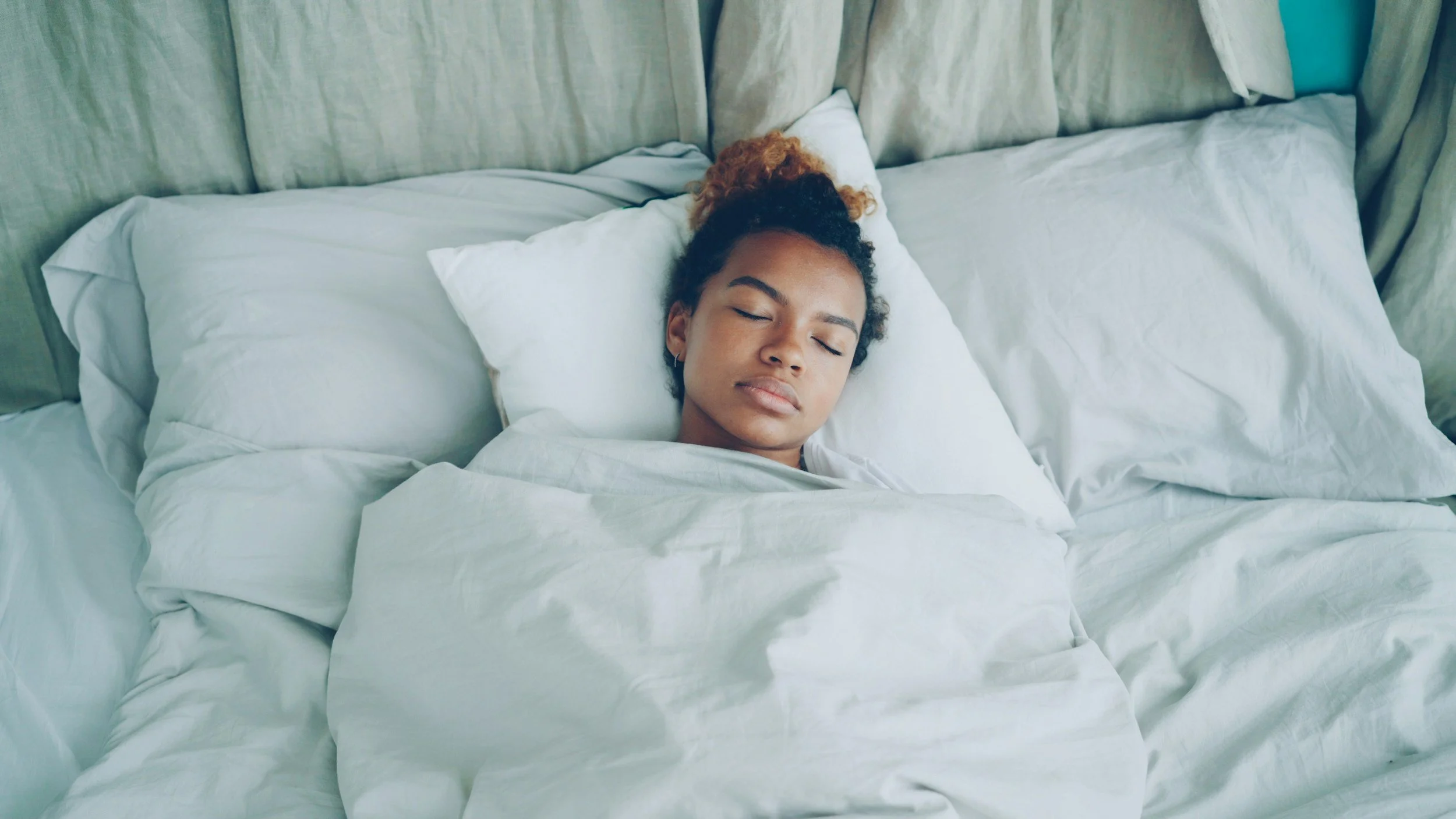 A teenage girl with curly hair, lying on her back in bed with eyes closed, surrounded by white sheets and pillows.
