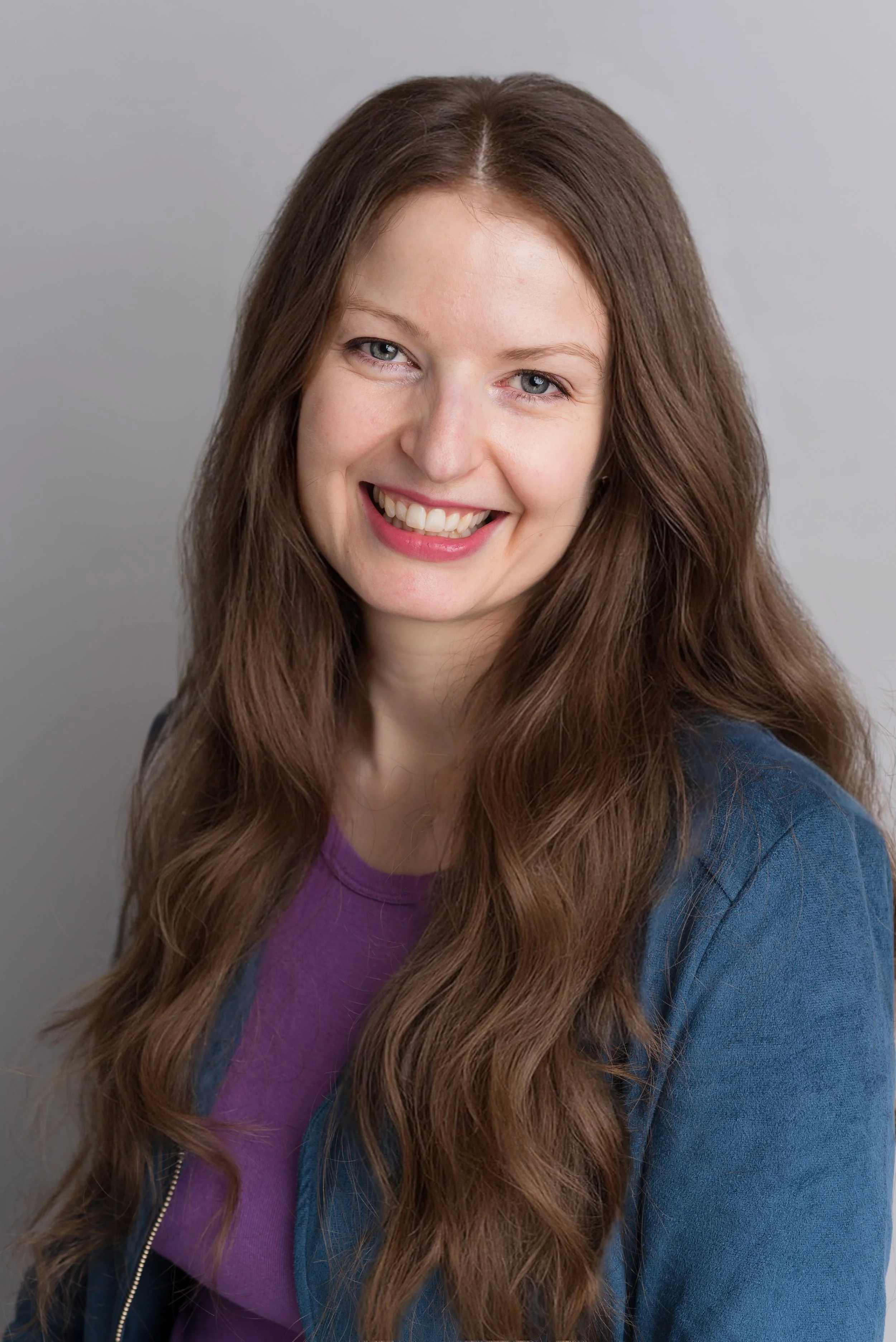 A young woman with long, wavy brown hair, blue eyes, and a bright smile wearing a purple top and a blue jacket against a plain grey background.