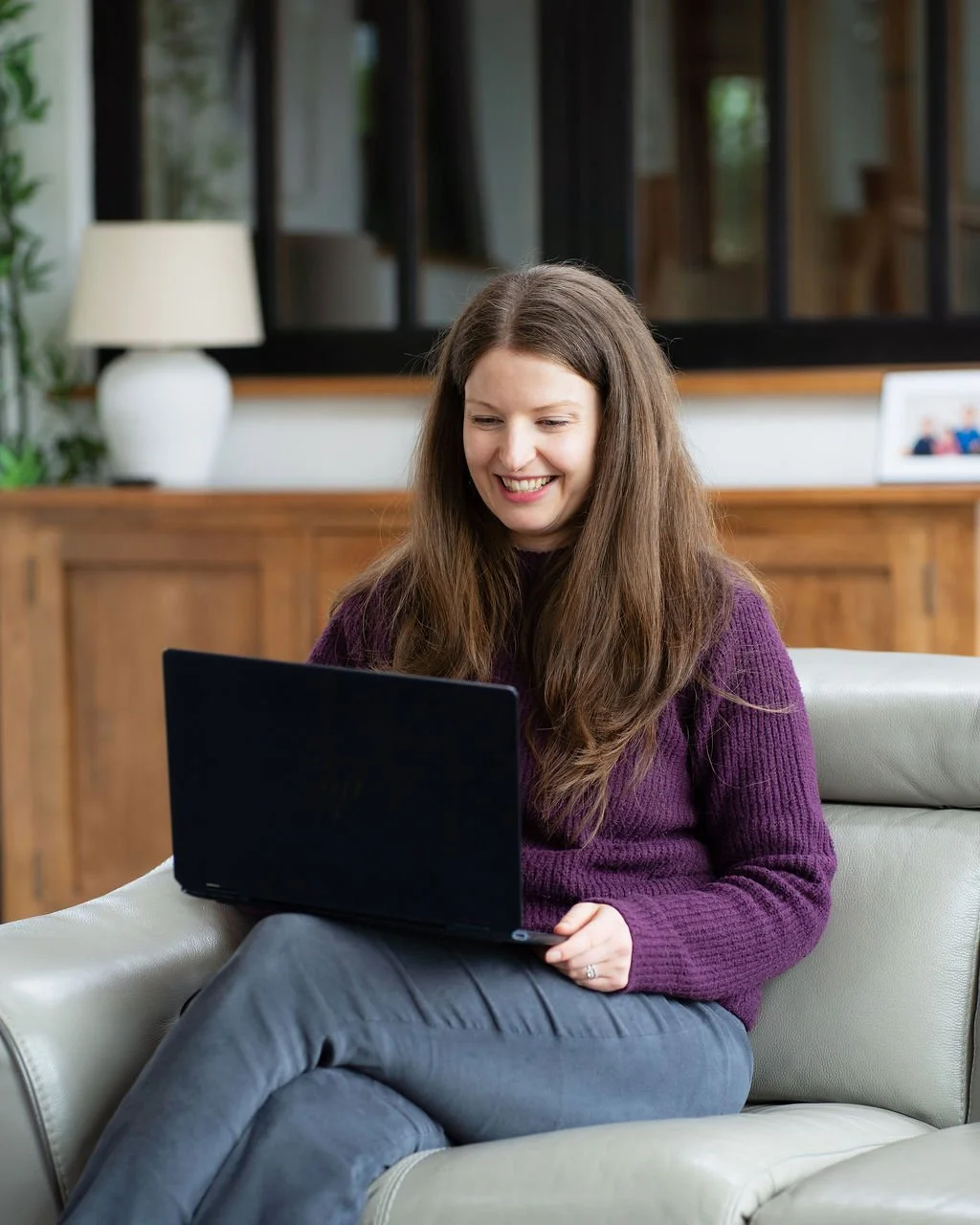 A young woman with long brown hair, wearing a purple sweater and grey trousers, sitting on a light grey sofa in a living room, smiling and looking at a black laptop on her lap.