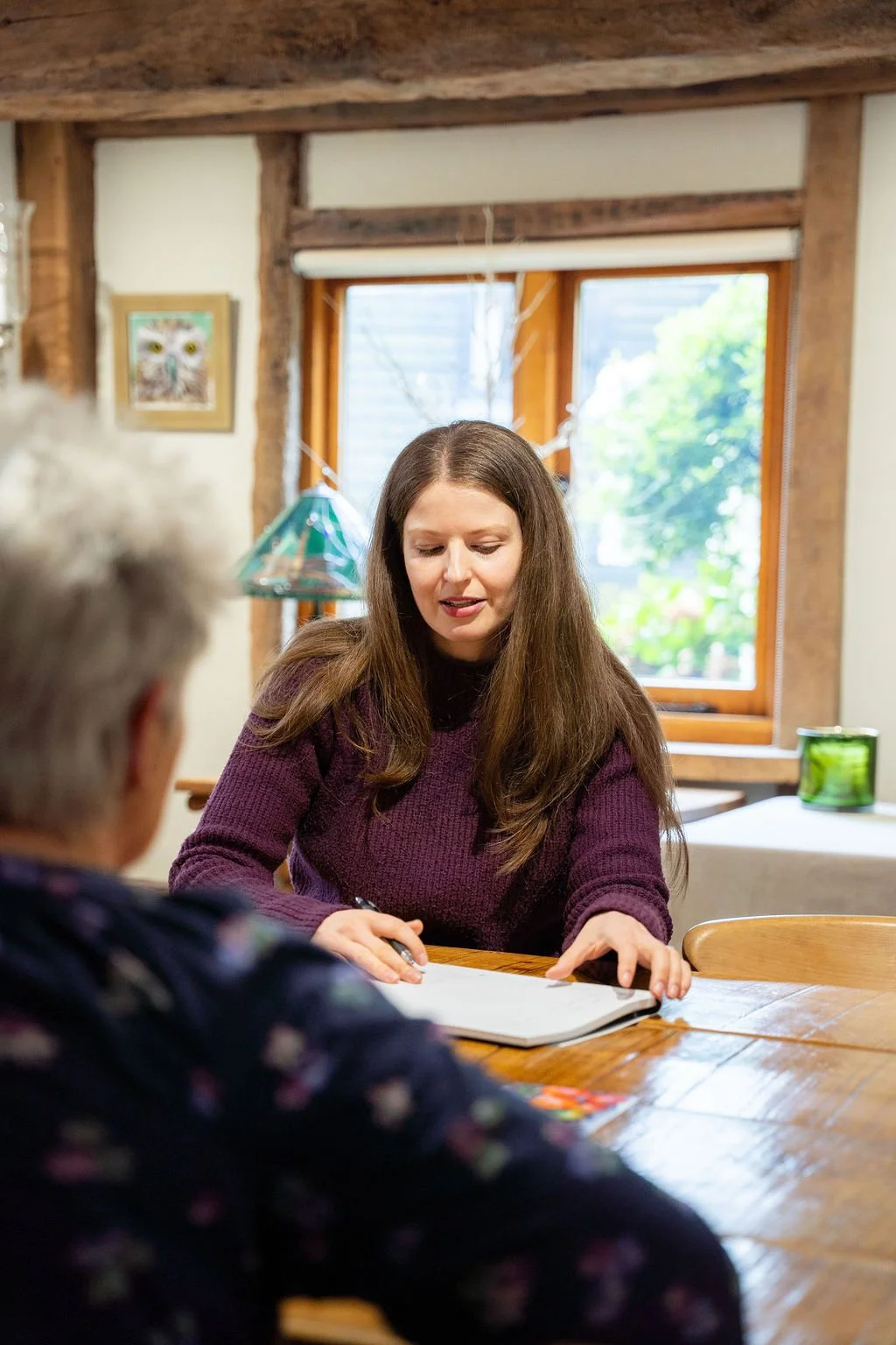 A woman with long brown hair sitting at a wooden table, engaged in a conversation with a grey-haired woman, in a cozy room with wooden beams and large windows showing greenery outside.