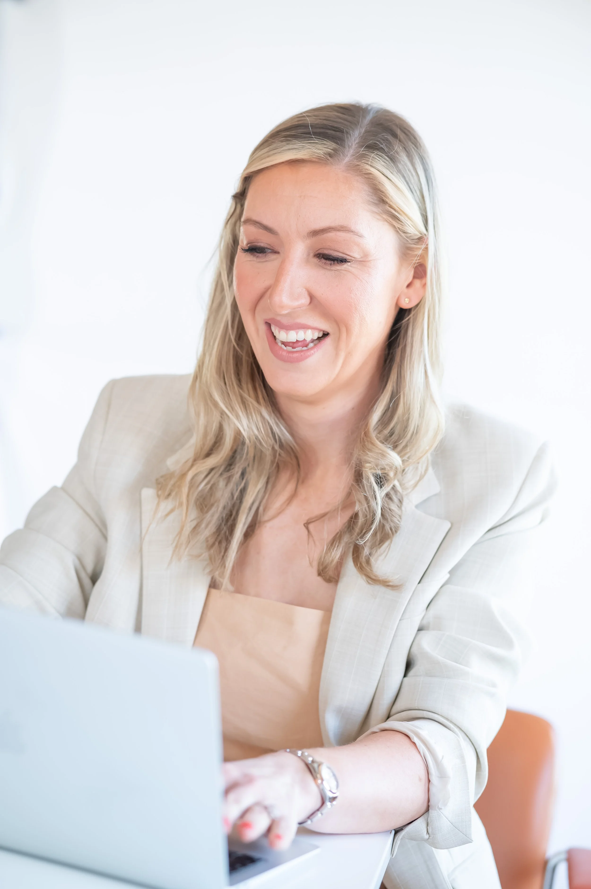 A woman with blonde hair smiling and working on a laptop in a bright, modern office.
