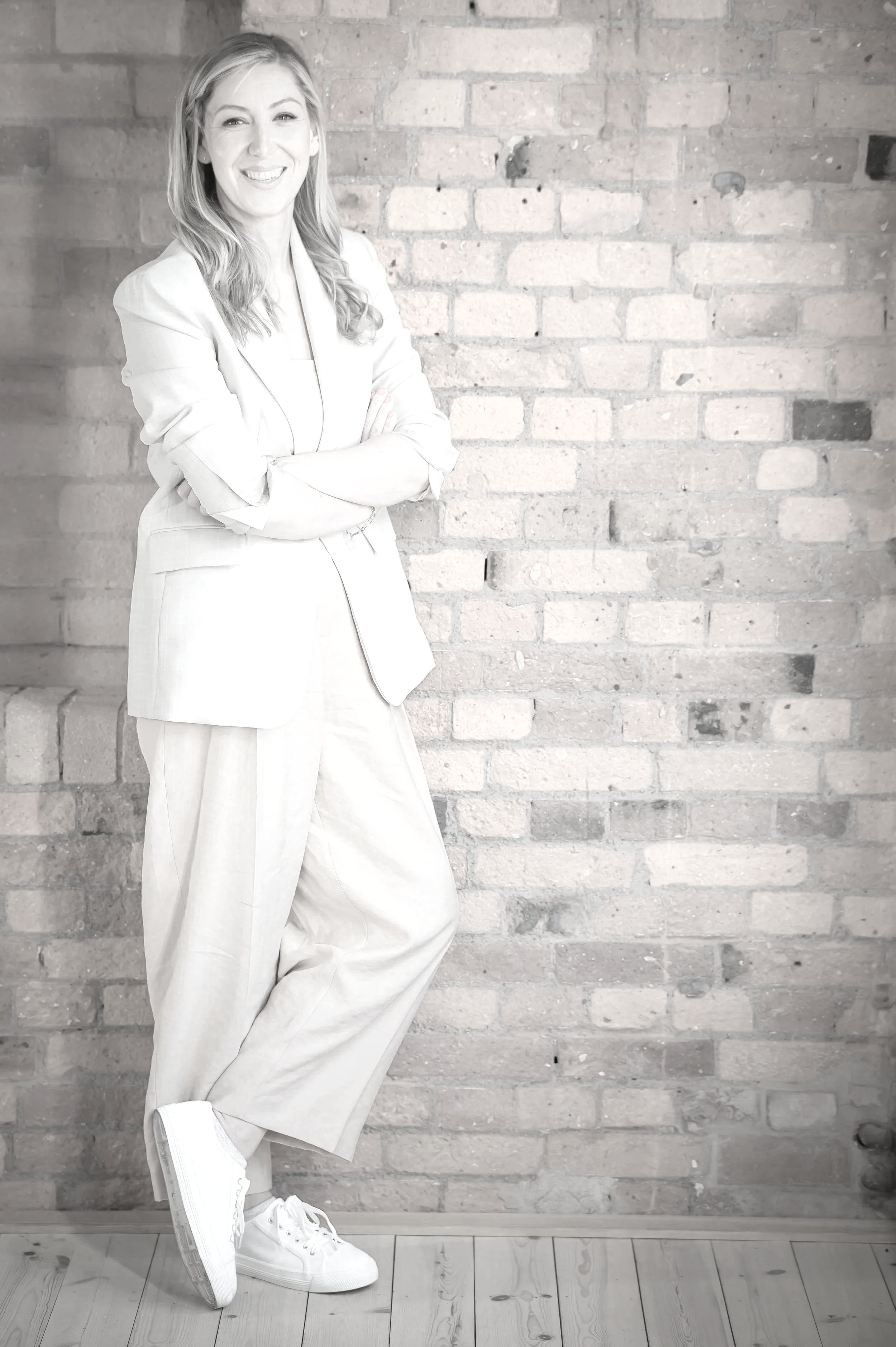 A woman standing against a brick wall, smiling with arms crossed, wearing a light blazer and pants with sneakers.