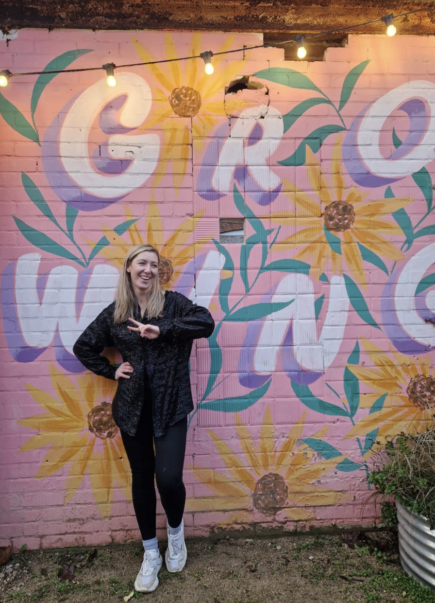 A smiling woman with blonde hair standing in front of a pink brick wall painted with large, colorful flowers and the words 'CRO' and 'WLO.' There are string lights hanging above her, and she is making a peace sign with her right hand. She is dressed in a black sparkly jacket, black pants, and white sneakers.
