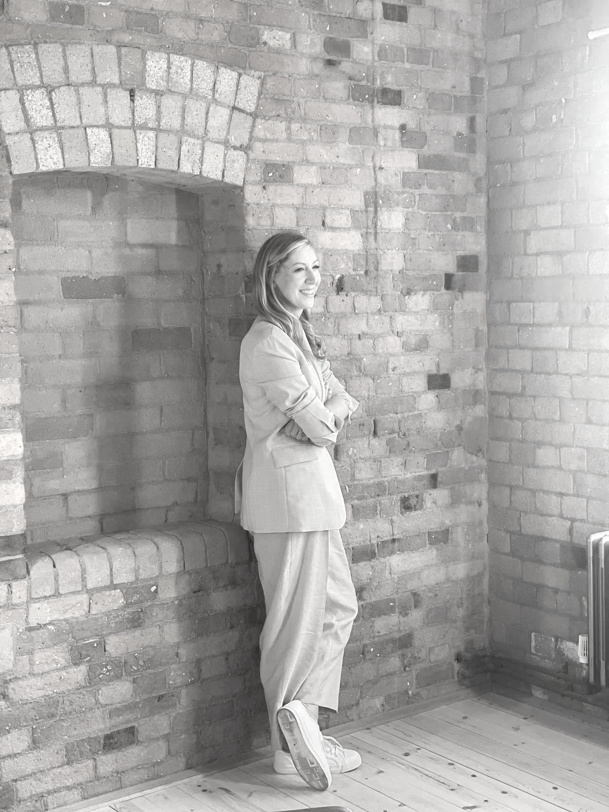 A woman smiling and standing with arms crossed in front of a brick wall, wearing a blazer and loose pants in a room with wooden flooring and a radiator.