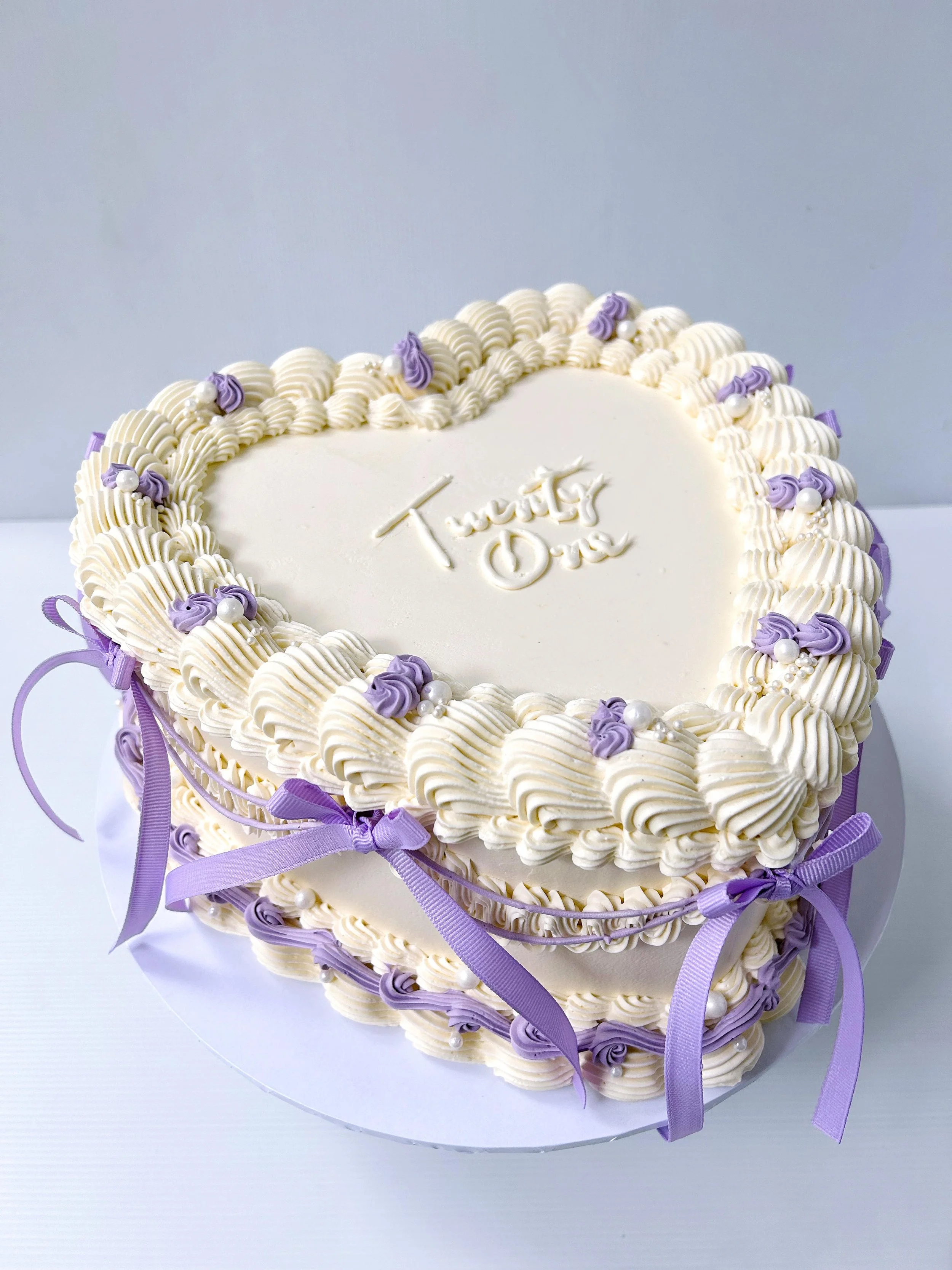 A heart-shaped birthday cake decorated with white and purple icing, ribbons, and pearls, with 'Twenty One' written on top.