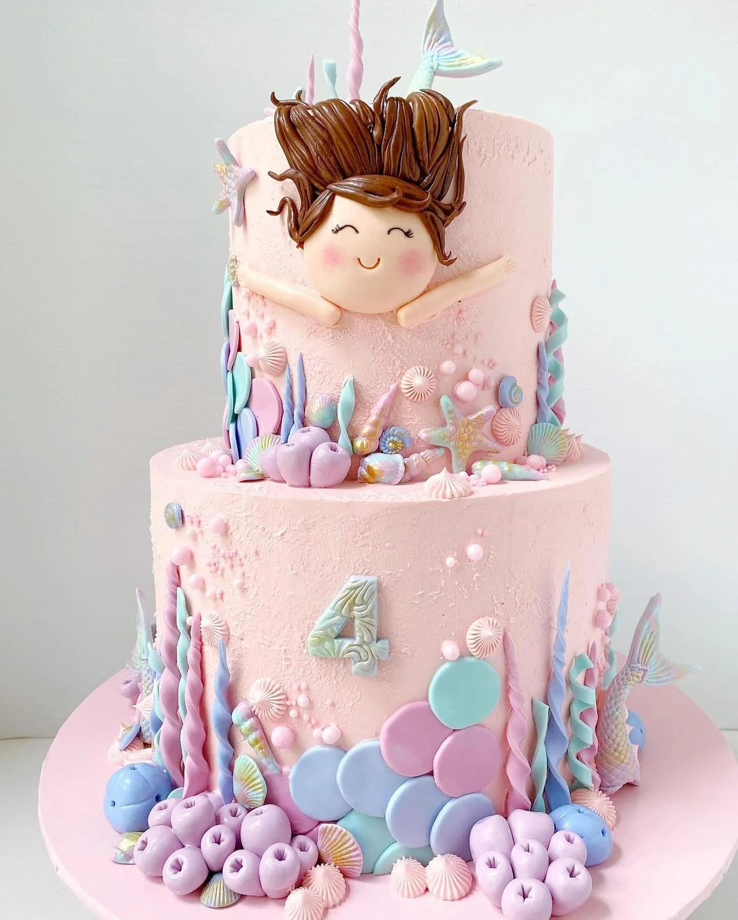A two-tier pink birthday cake decorated with pastel seashells, starfish, and cherries, featuring a doll face with animated hair and arms as the cake topper, with the number 4 on the lower tier.