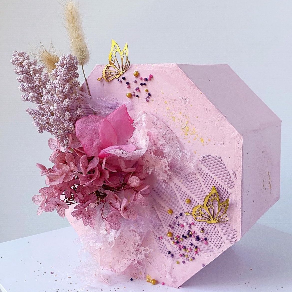 A pink hexagonal gift box decorated with pink flowers, dried plants, and gold butterfly ornaments, with colorful beads and gold accents on the surface.