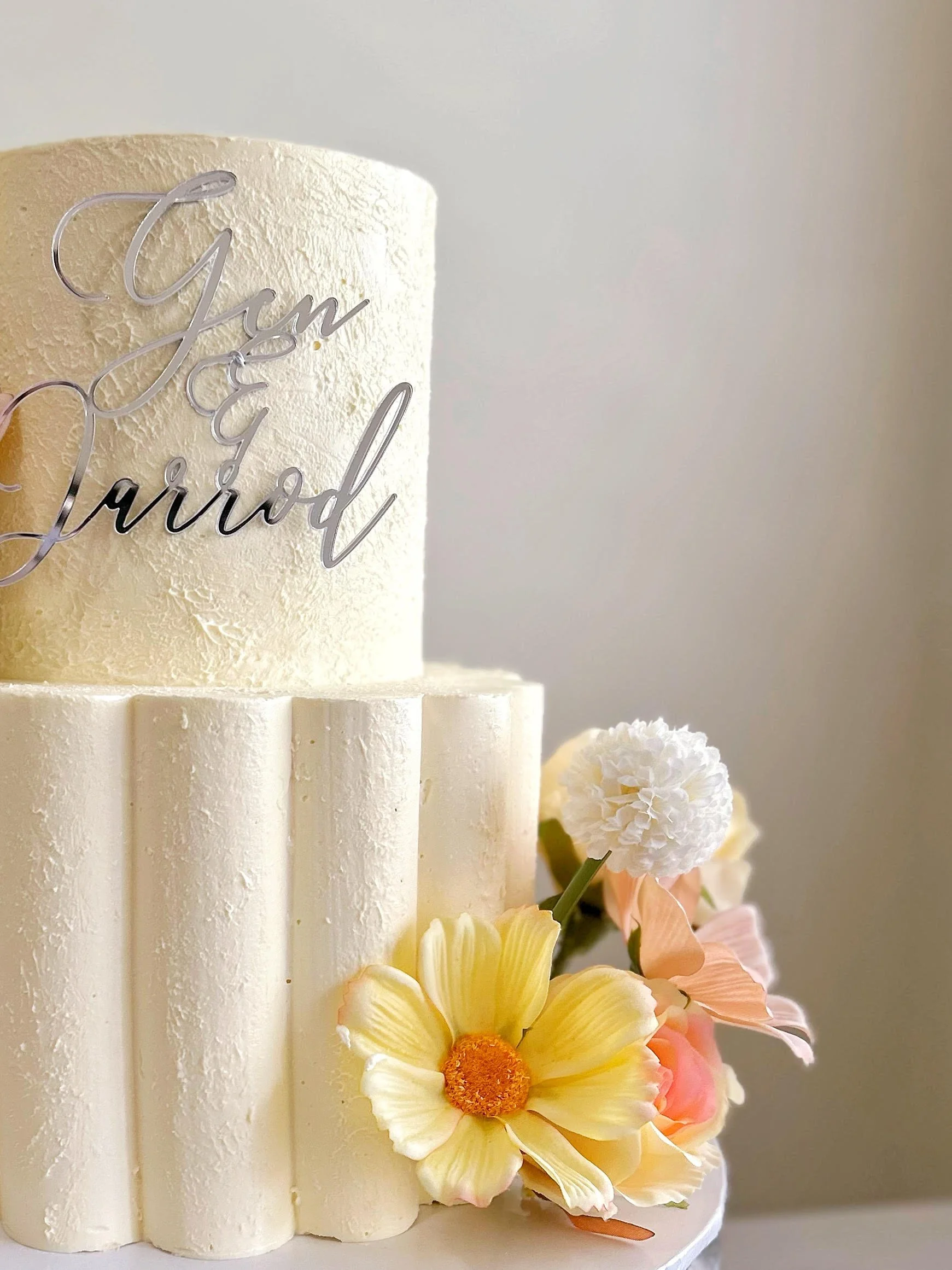 Close-up of a white wedding cake decorated with fresh flowers and a silver cake topper that says 'I'm so proud'.