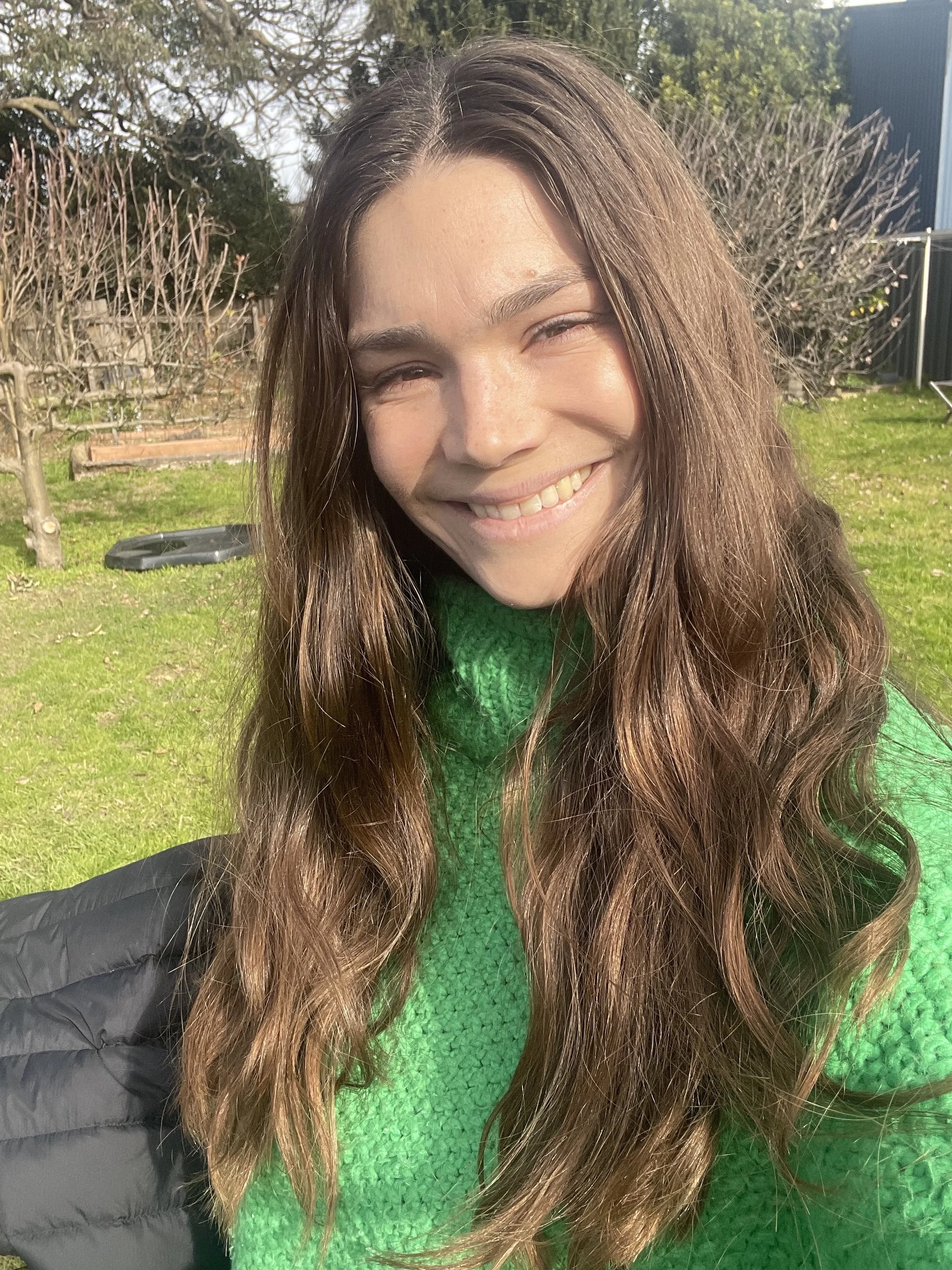 Young woman with long brown hair smiling outdoors in sunlight, wearing a green sweater and black jacket, with a lush garden background.