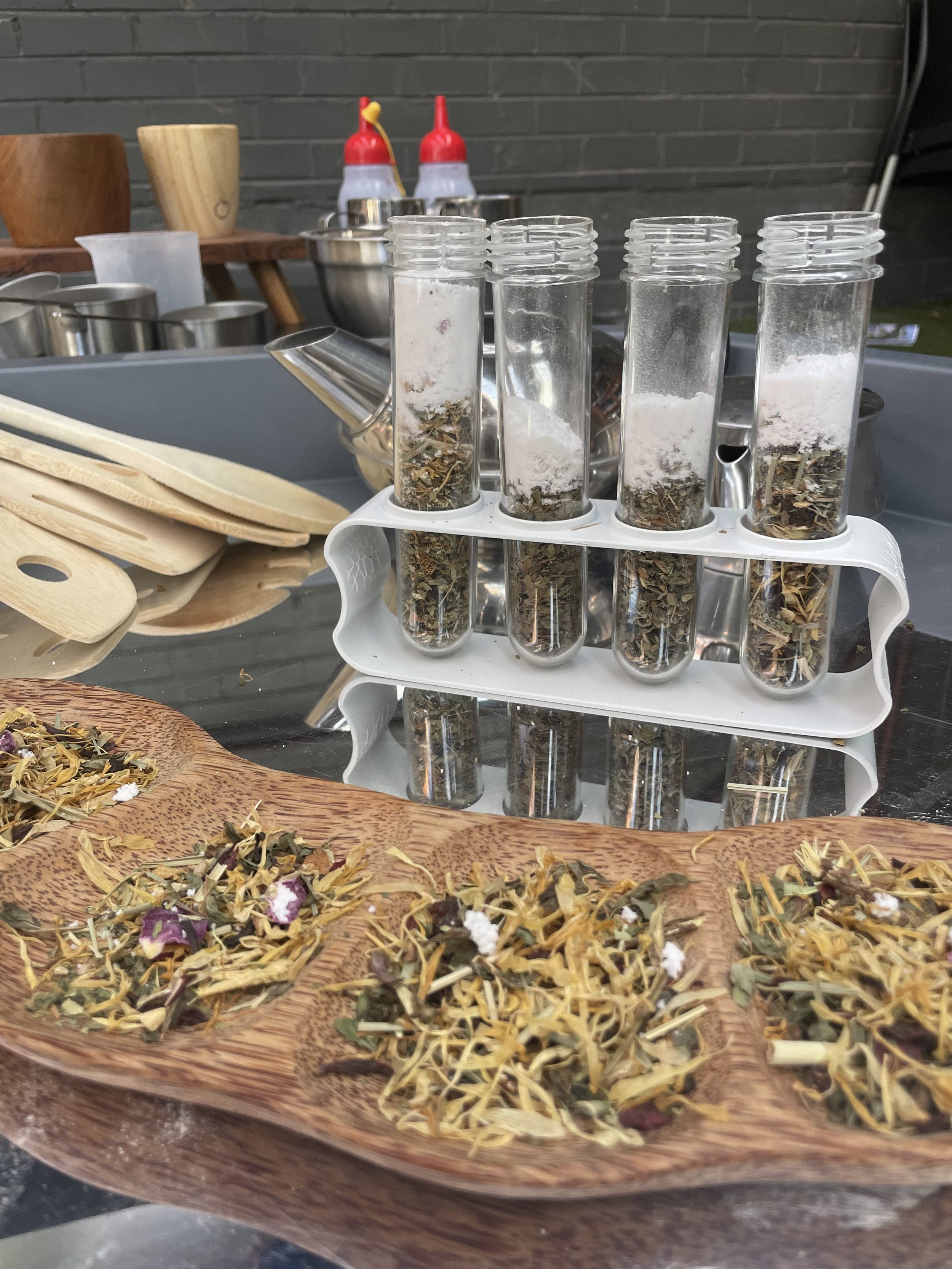 Test tubes filled with dried herbs on a white rack with dried herbs on a wooden tray in front, kitchen utensils and condiments in the background.