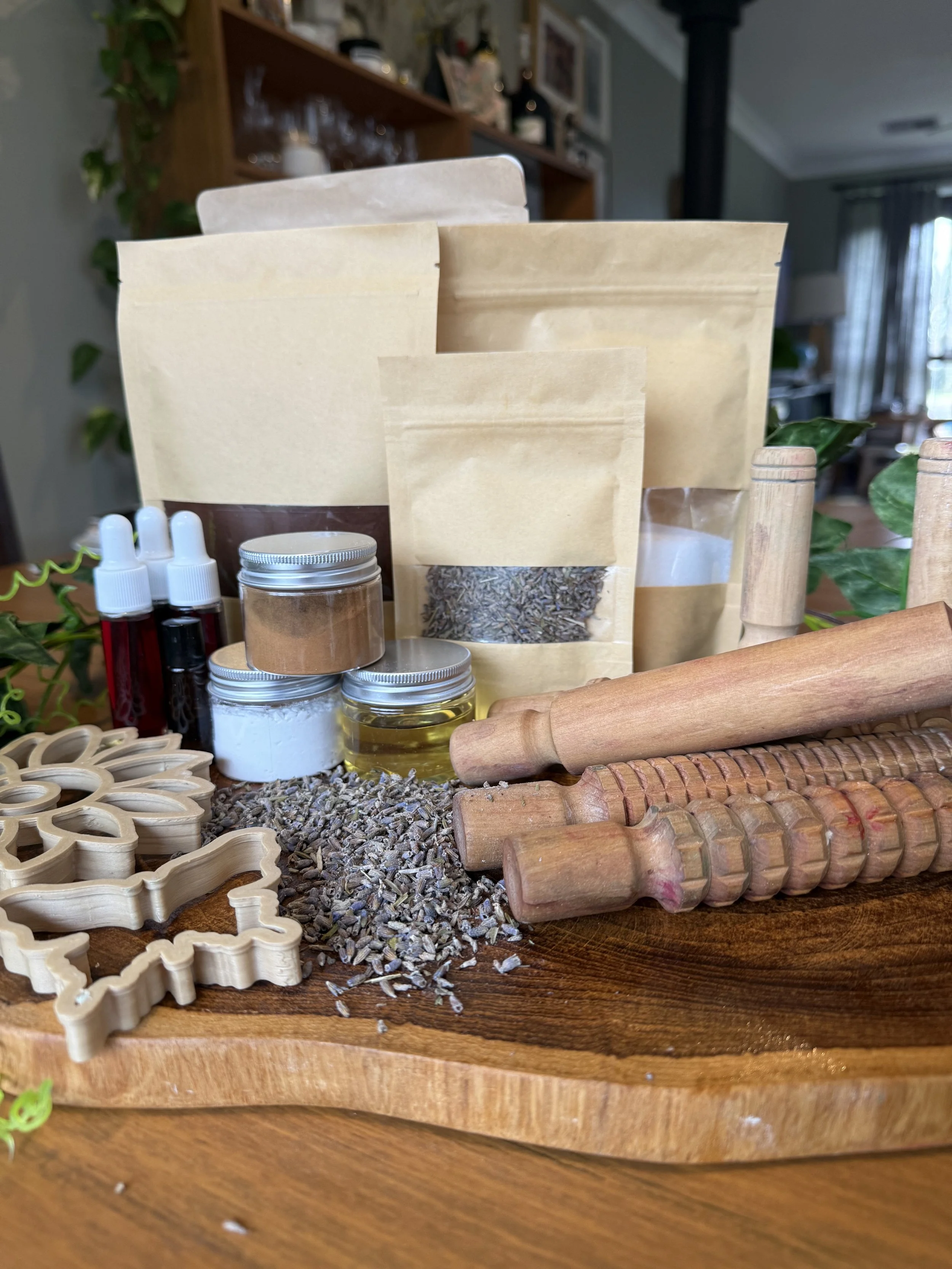 Assorted kitchen or baking supplies on a wooden tray, including rolled pins, small jars of ingredients, dried herbs or lavender, bottles with droppers, and paper bags, with a blurred living room background.