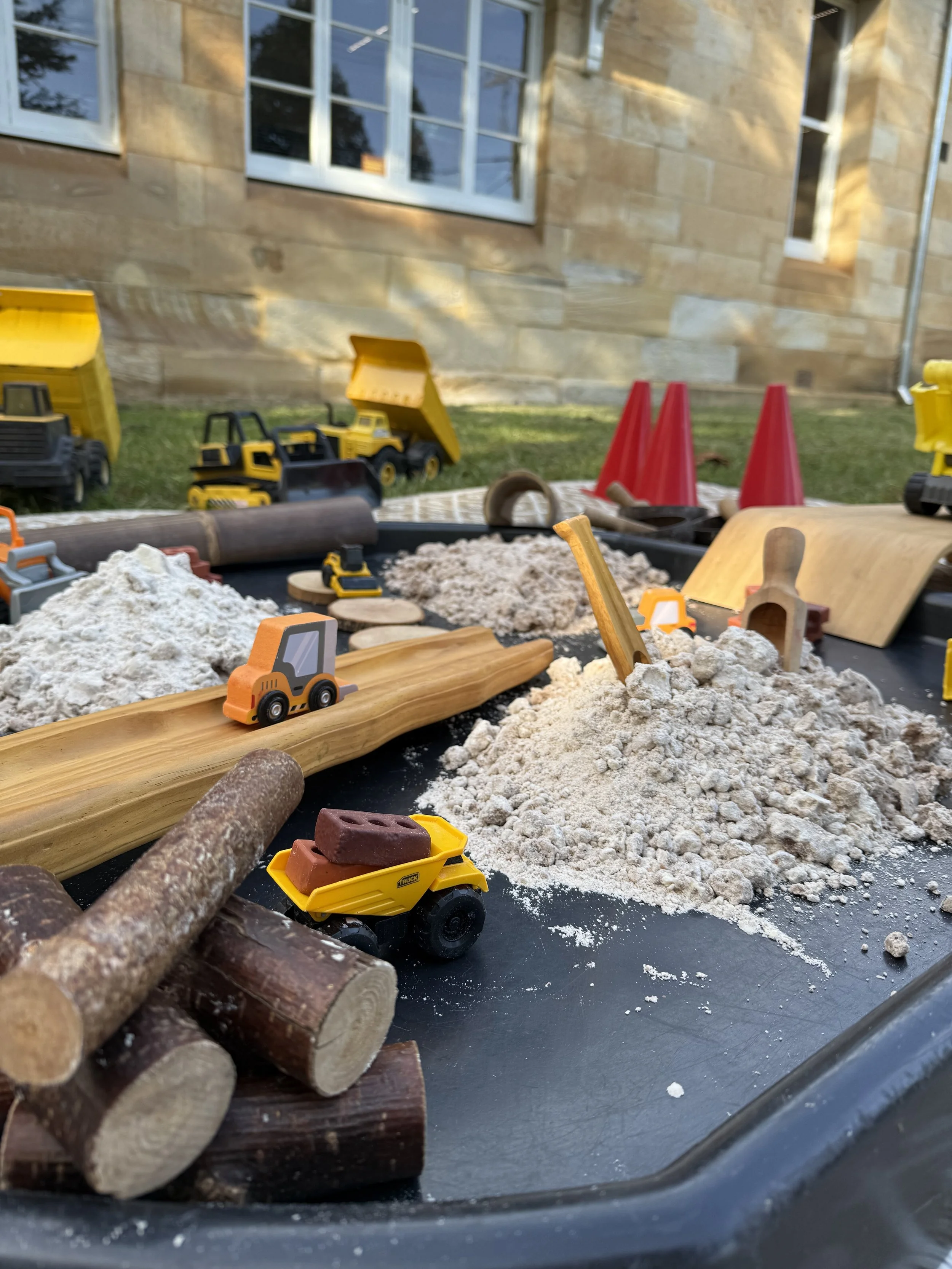 Children's sand and water table set up outdoors with toy construction vehicles, sand, rocks, cones, and wood logs on a sunny day.
