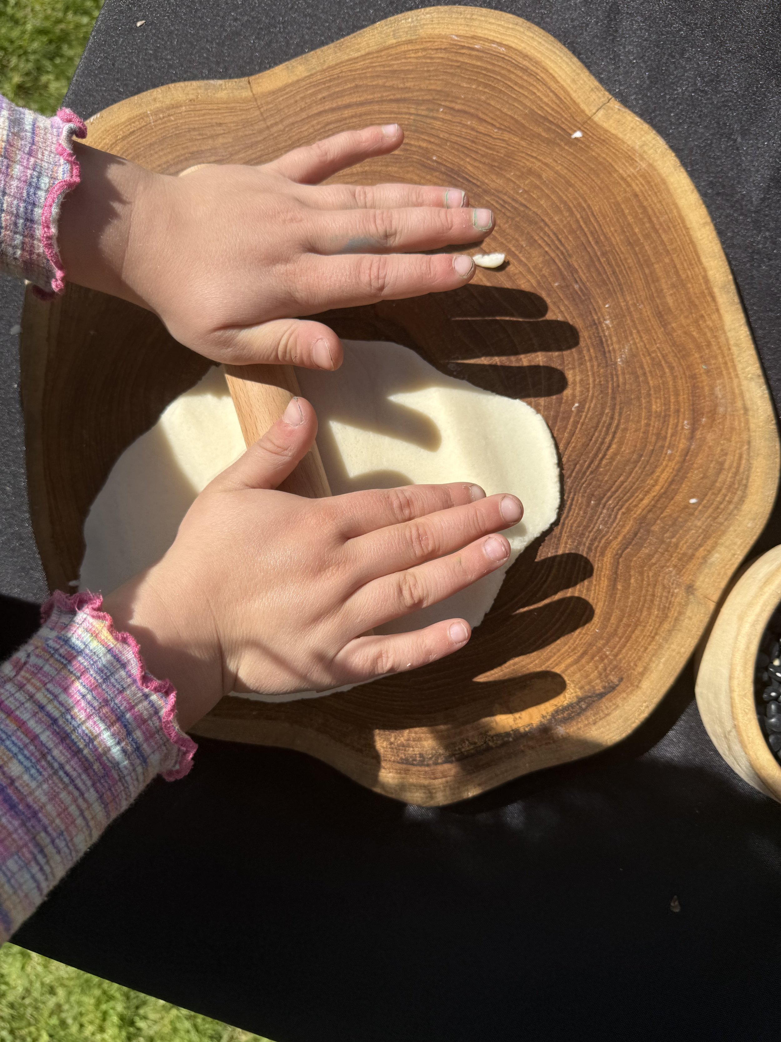 Child rolling out dough with a rolling pin on a wooden board outdoors.