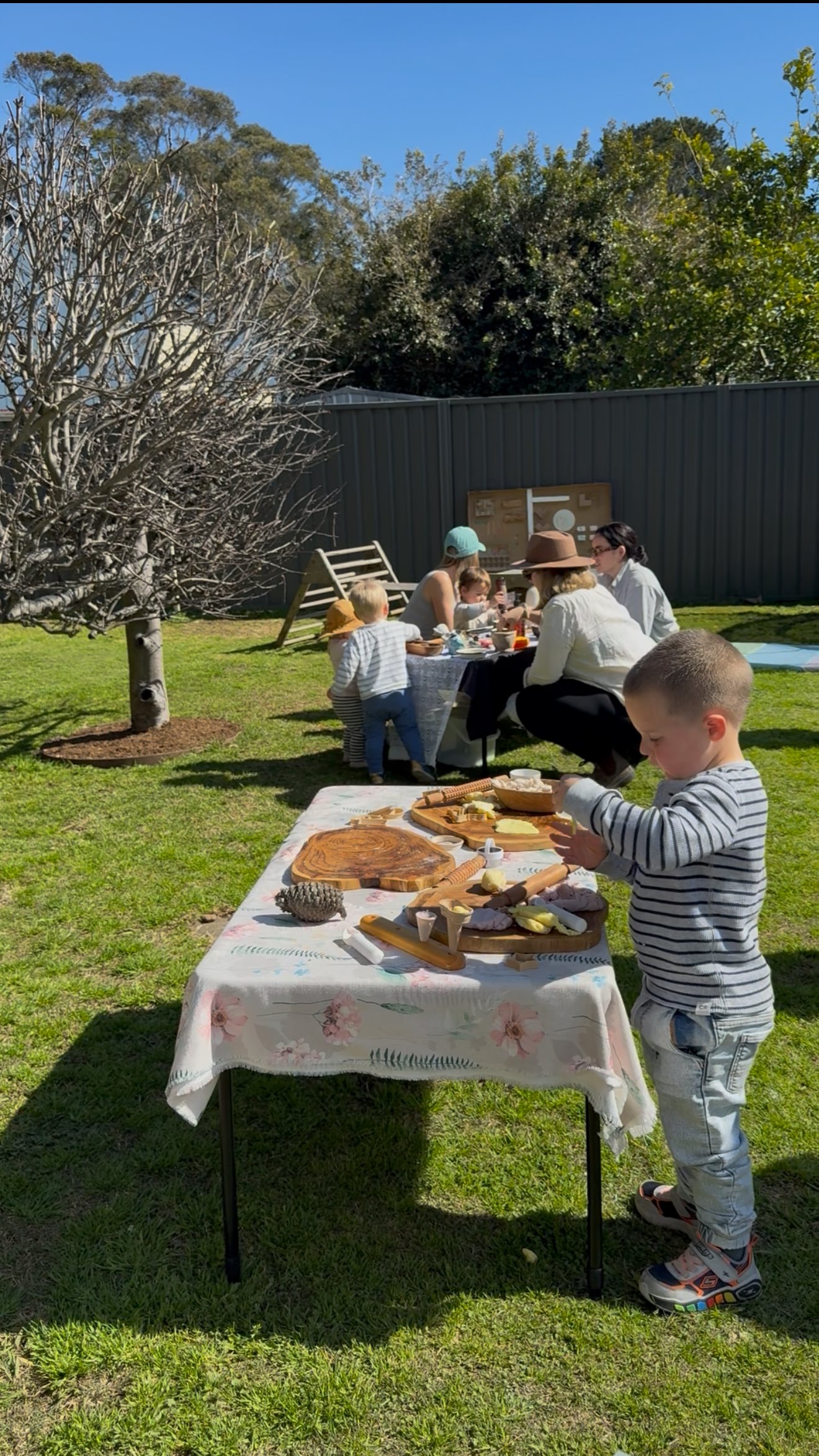 Children and adults having a picnic in a backyard on a sunny day. A young boy in striped shirt reaches for food on a table with baked goods. Two tables are set with snacks and desserts, surrounded by grass, a tree, and a dark fence.