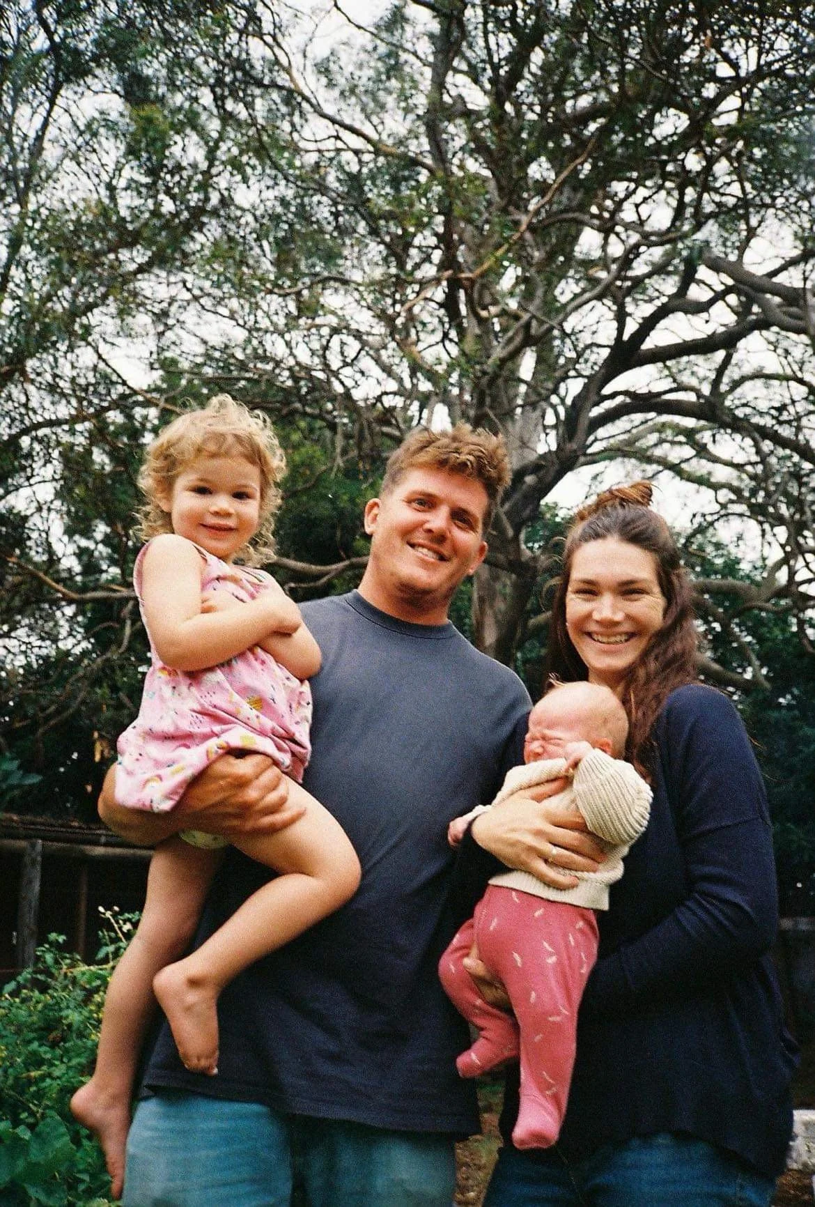 A family of four outdoors with a large tree in the background. The father is holding a little girl in a pink dress, and the mother is holding a baby in a cream sweater and pink pants. Everyone is smiling.