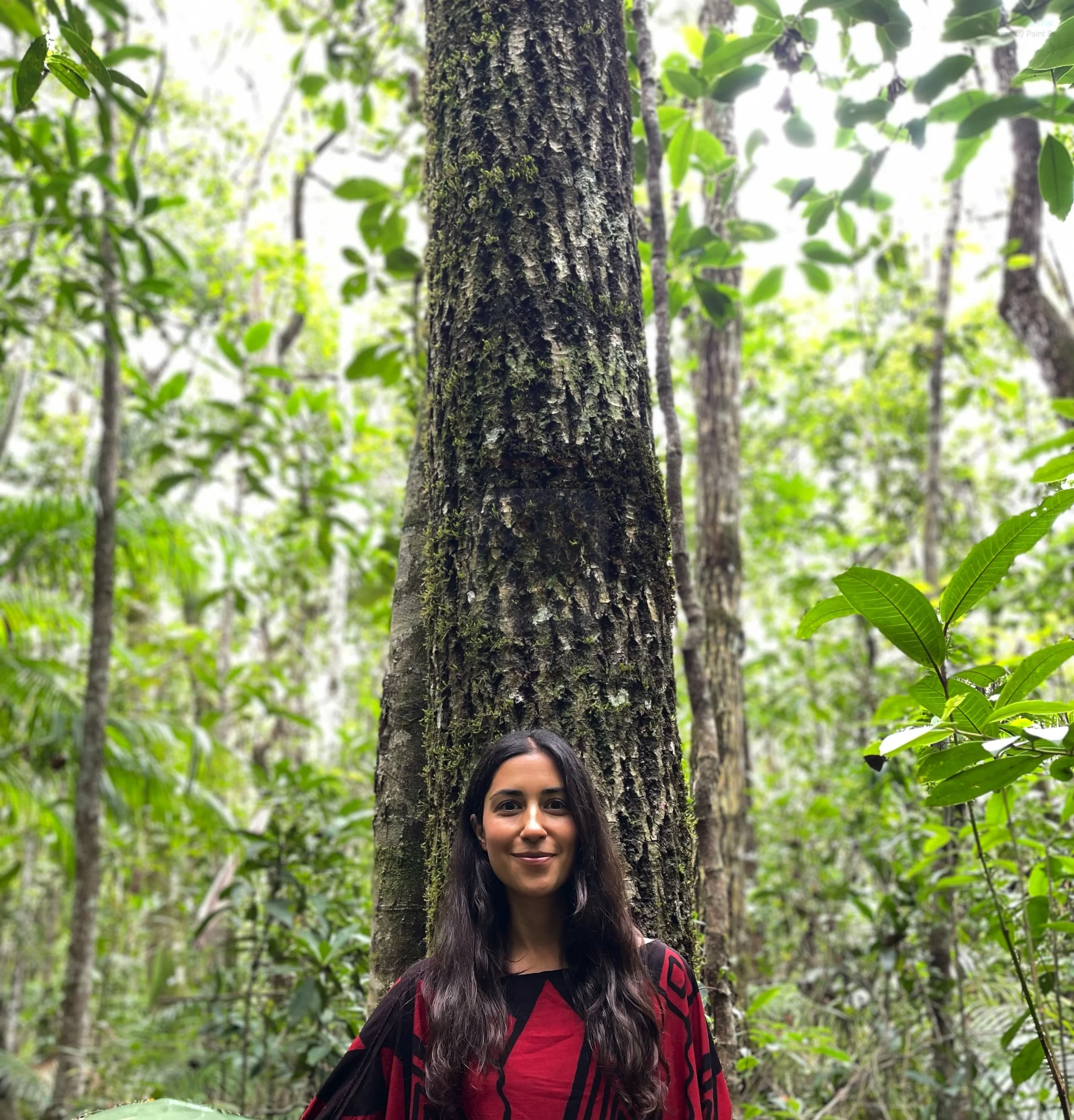 A woman with long dark hair smiling in a dense green forest, standing in front of a tall tree trunk.