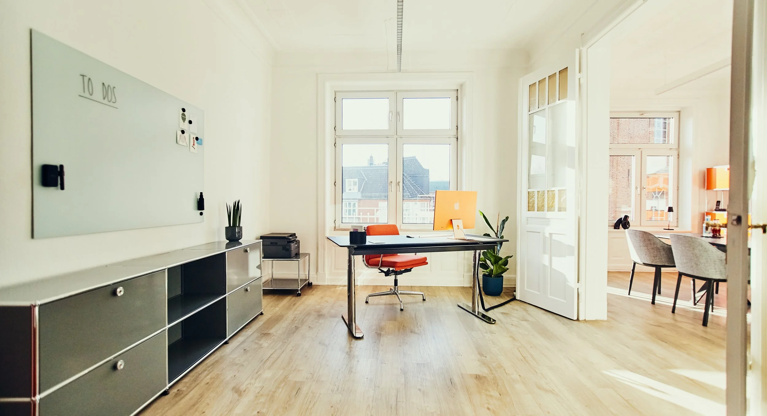 Helles, modernes Büro mit großem Fenster, Schreibtisch mit orange Stuhl, Pflanzen, Whiteboard, in einem angrenzenden Raum another Arbeitsbereich mit Schreibtisch und Stühlen.