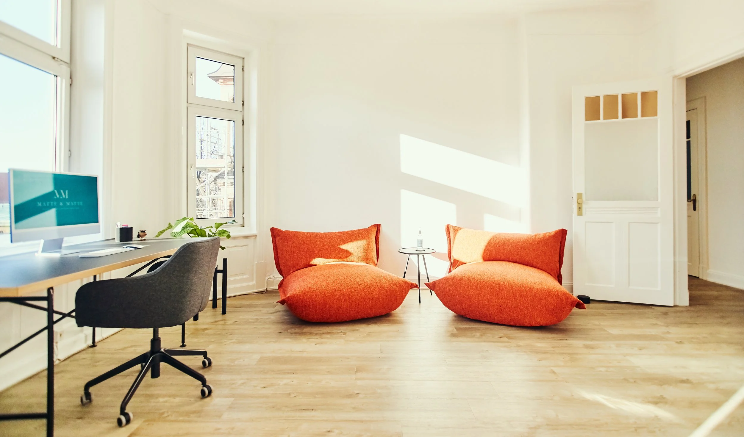 Helles, modernes Wohnzimmer mit zwei großen, orangefarbenen Sesseln, einem kleinen Beistelltisch mit Wasserflasche, einem Schreibtisch mit Computer und einem Stuhl, mehrere große Fenster lassen viel Sonnenlicht herein.