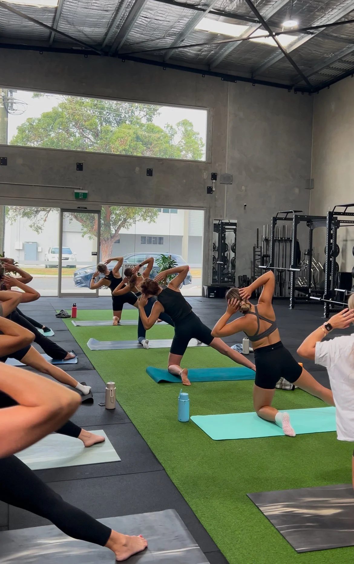 A group of people in a gym performing a fitness class, doing stretches on mats on artificial grass.