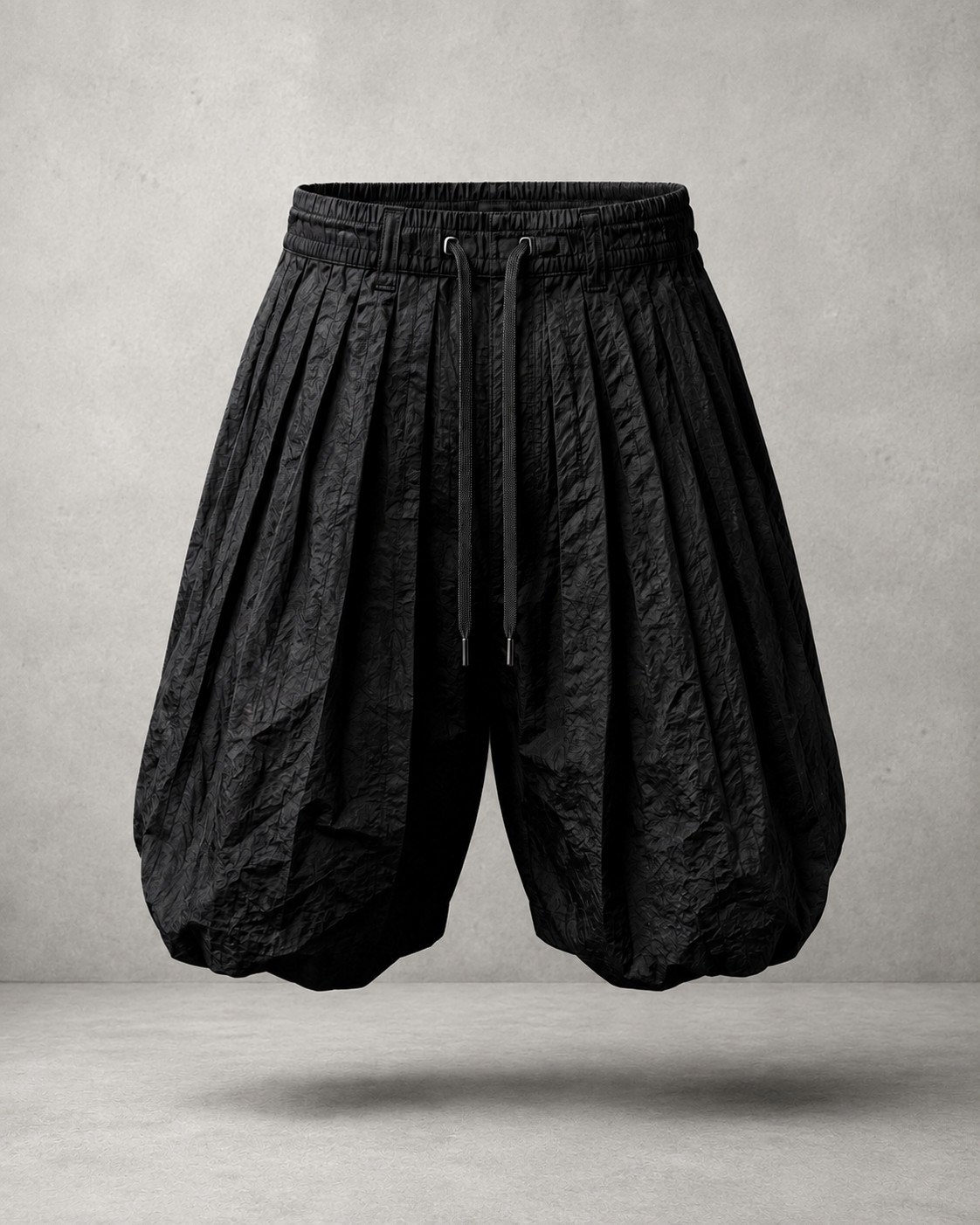 Black, textured, loose-fitting, pleated shorts or pants with a drawstring waistband, suspended in mid-air against a gray background.