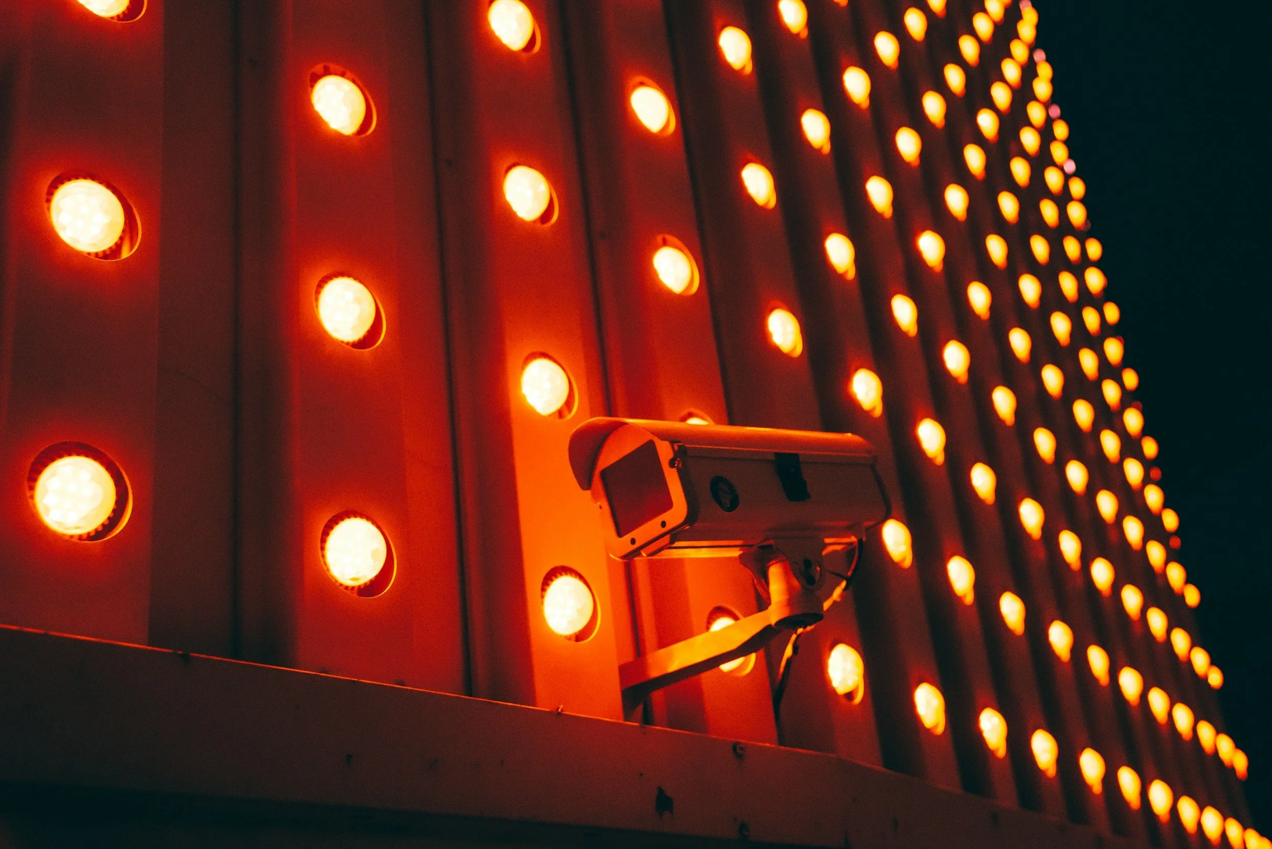 A surveillance camera mounted on a building surrounded by bright orange circular lights, creating a visually striking pattern at night.