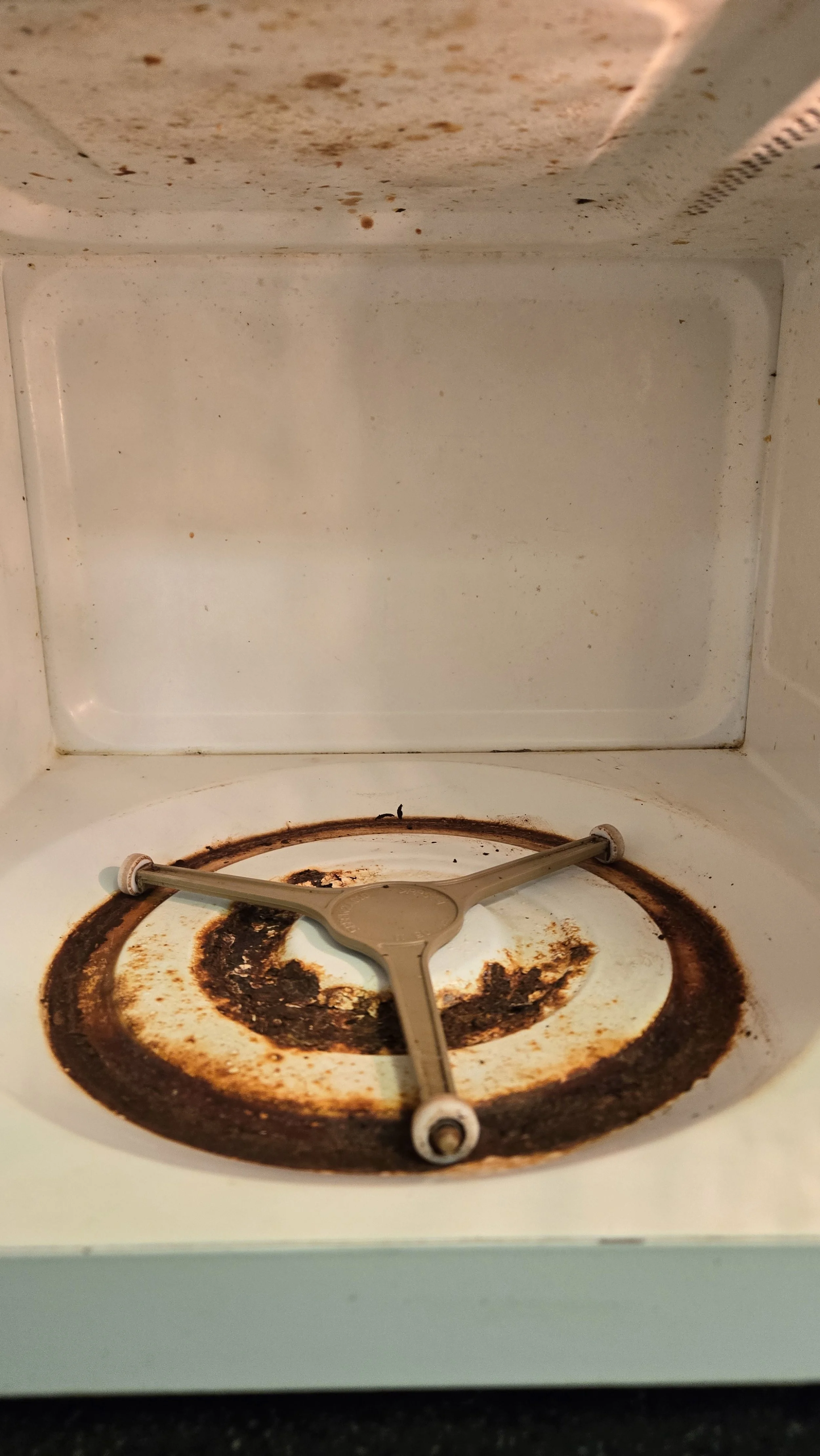 Inside a microwave with a dirty, rusty glass turntable and a broken Y-shaped metal support.