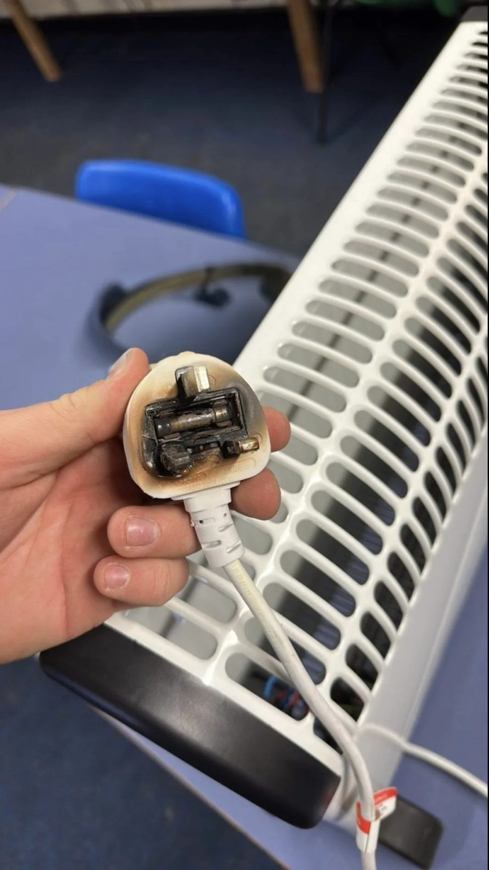 A hand holding a power adapter with burnt or damaged plug prongs in front of a space heater and a blue chair.