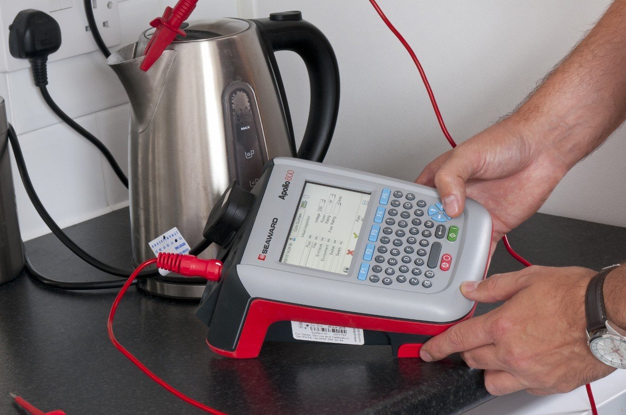 A person using a Seaworth Apollo 600 electrical testing device connected to a kettle on a kitchen countertop.