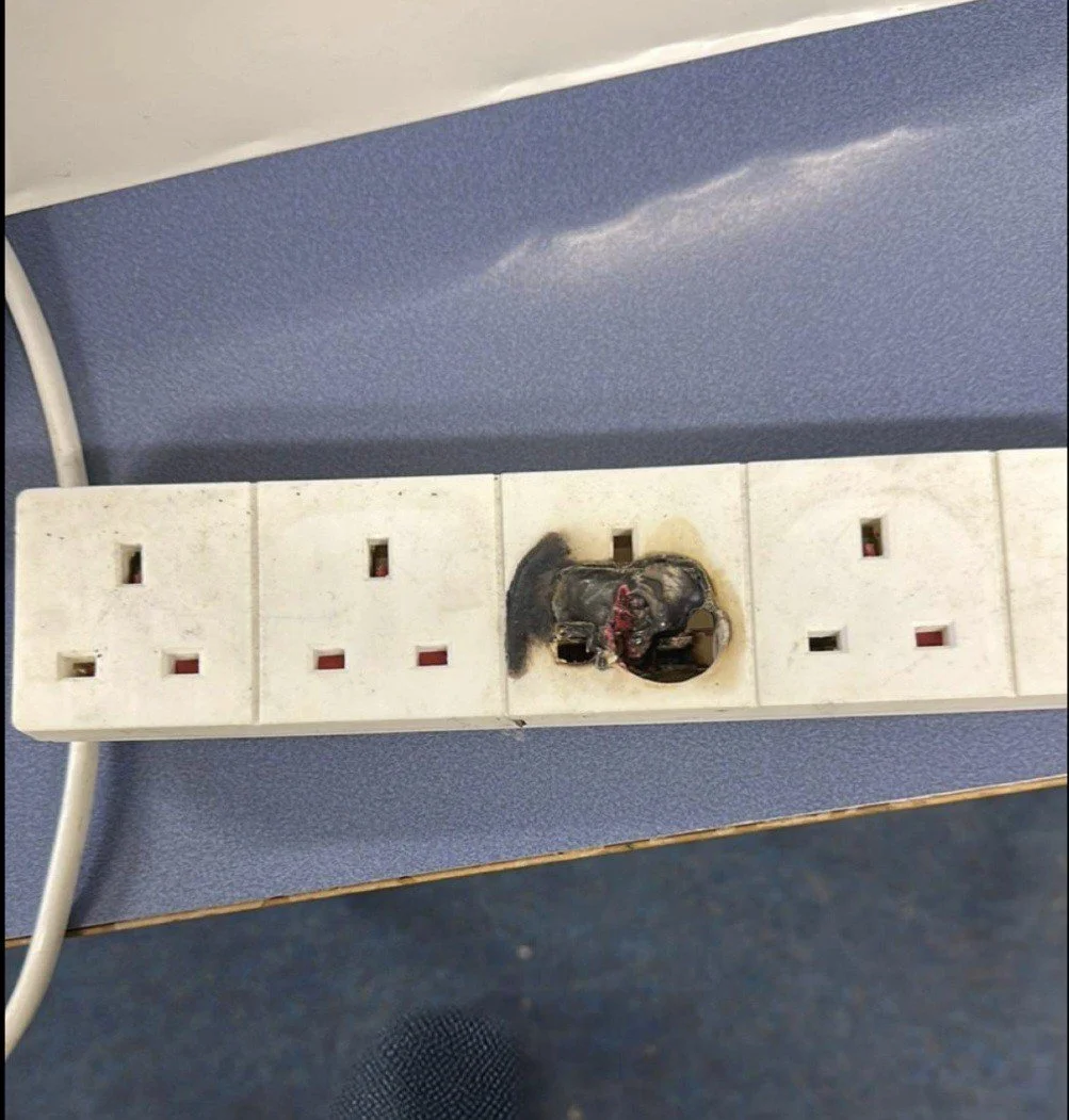 A power strip with multiple outlets, one of which has an electronic device that is burned and melted, with damage and charred residue visible around the socket.