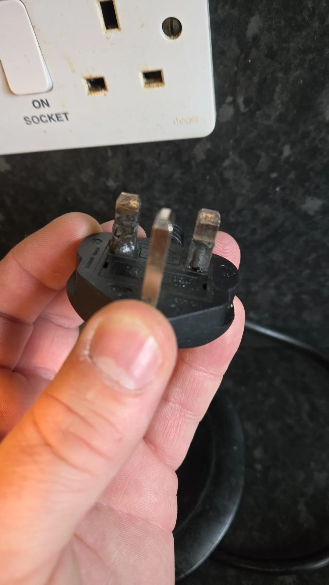 Hand holding a worn power plug near an electrical outlet on a black countertop.