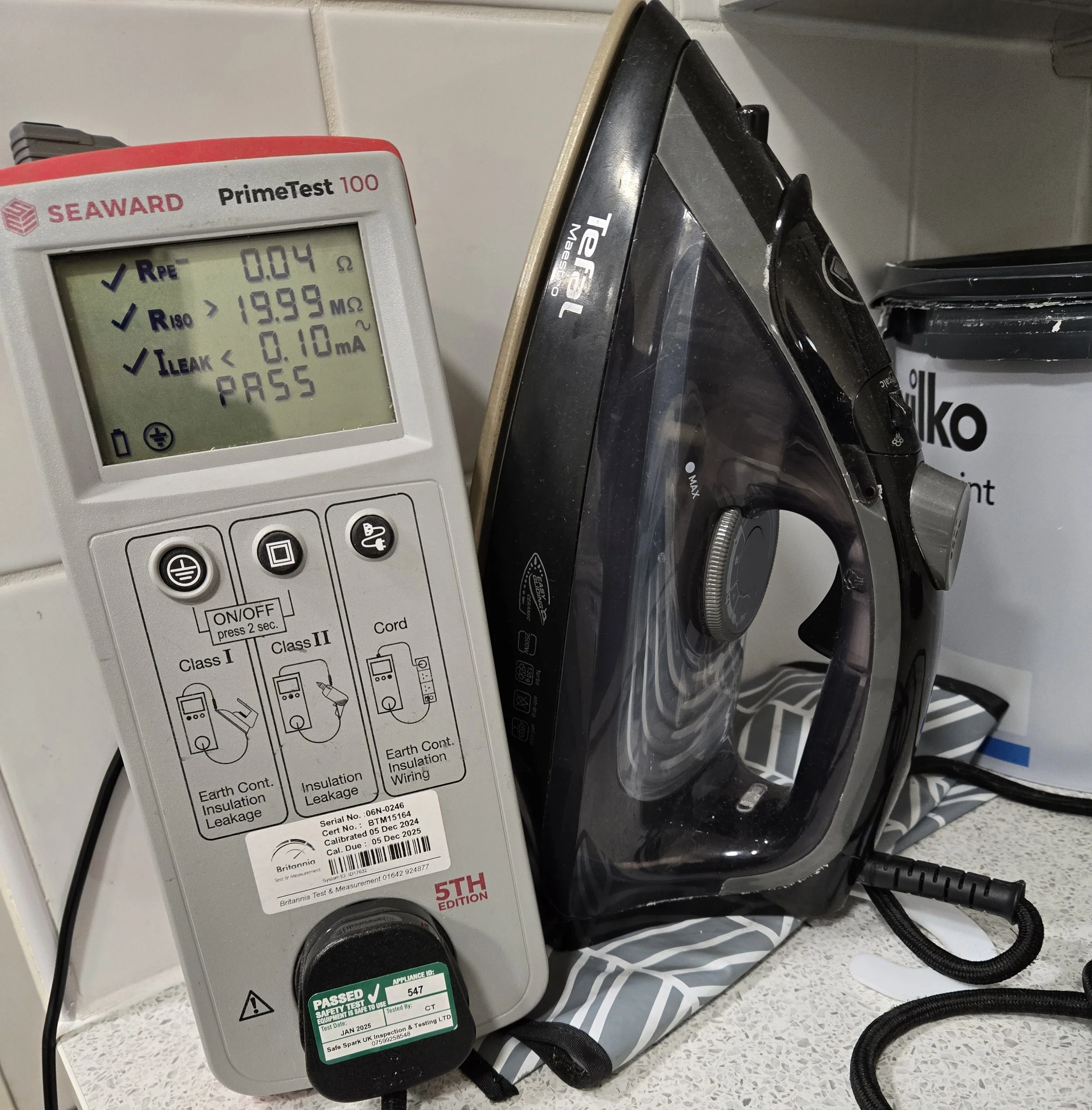 A multimeter displaying a reading of 0.04 ohms resistance, over 19.99 megaohms of resistance, and less than 0.10 milliamps leakage, with a passing status. The meter is next to a black cordless iron on a striped fabric mat, with a white container labeled 'SKO' in the background.