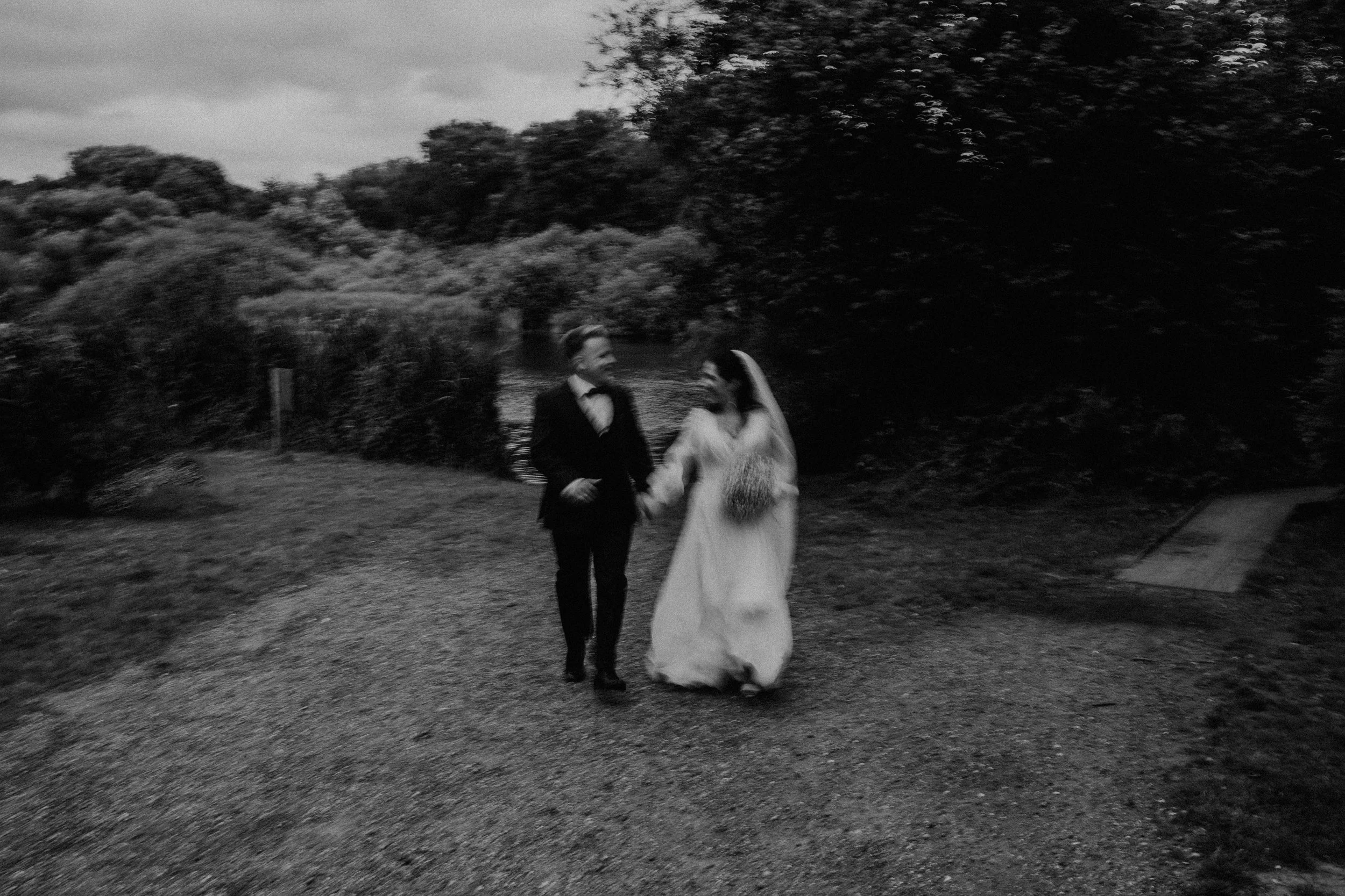 Zwei Menschen in formeller Kleidung, ein Mann in einem schwarzen Anzug und eine Frau in einem weißen Kleid, gehen Hand in Hand im Freien bei Dämmerung, umgeben von Bäumen und Natur.