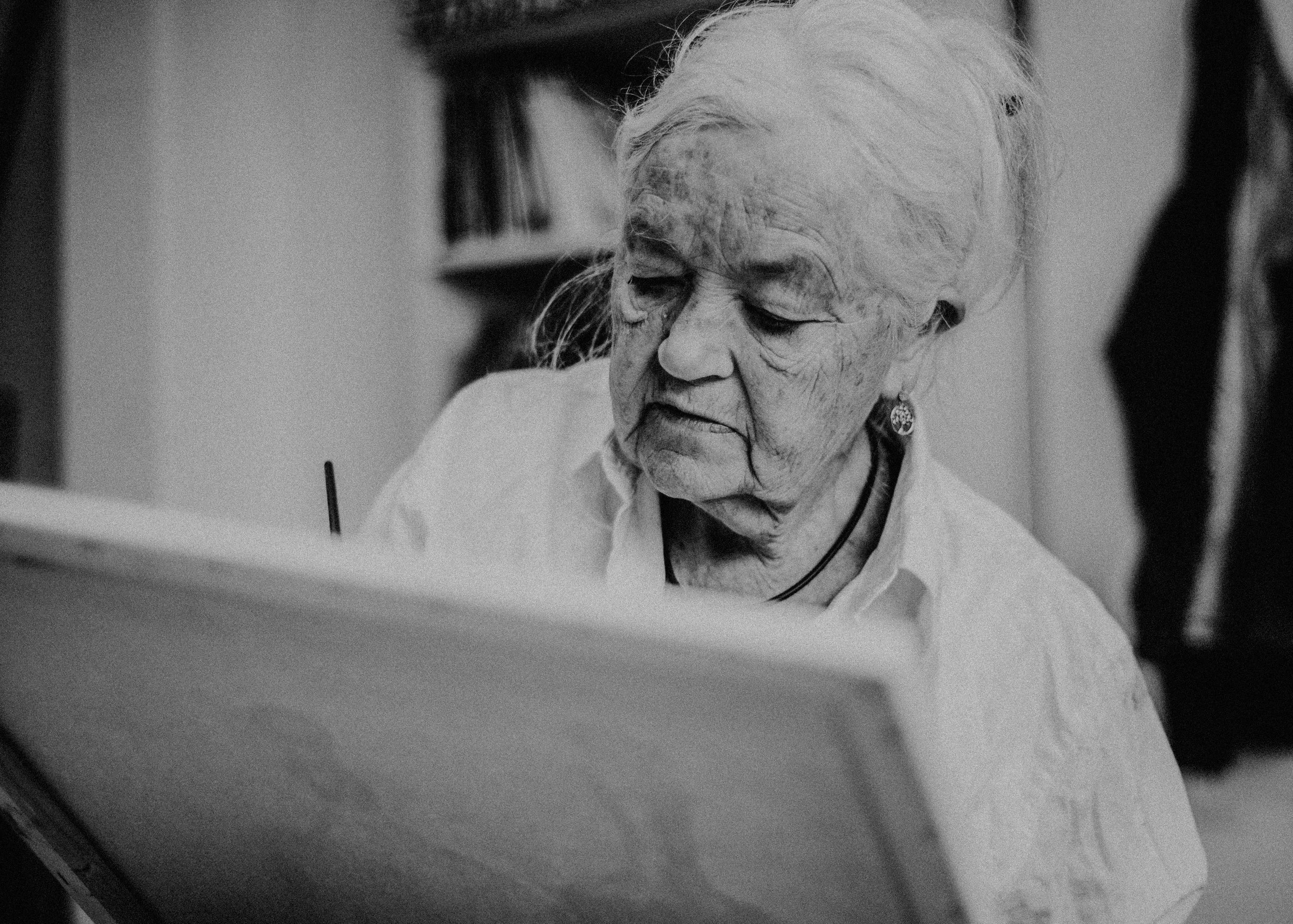 Eine ältere Frau mit weißen Haaren liest oder schreibt an einem Tisch.