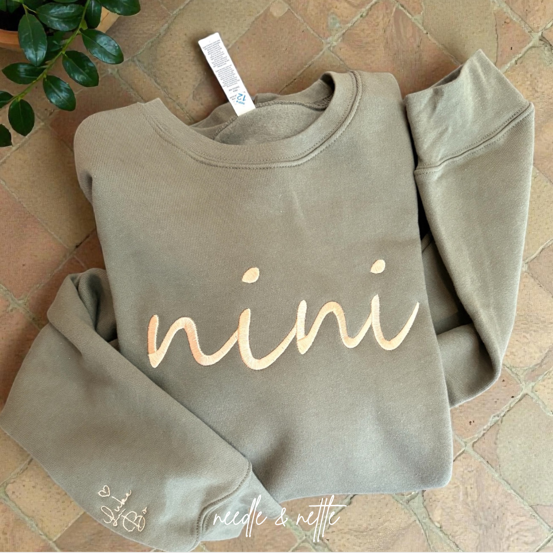 A taupe-colored sweatshirt with the word 'nini' embroidered in cursive on the front, laid on a tiled floor next to a green plant.
