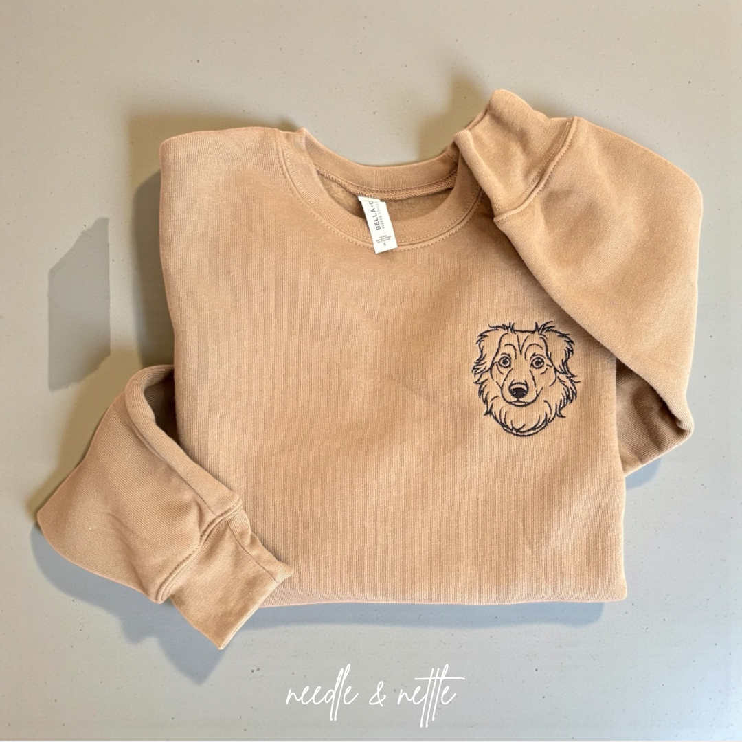 A beige sweatshirt with a dog face embroidered on the left chest, folded on a light-colored surface, with the words "needle & nettle" written below.