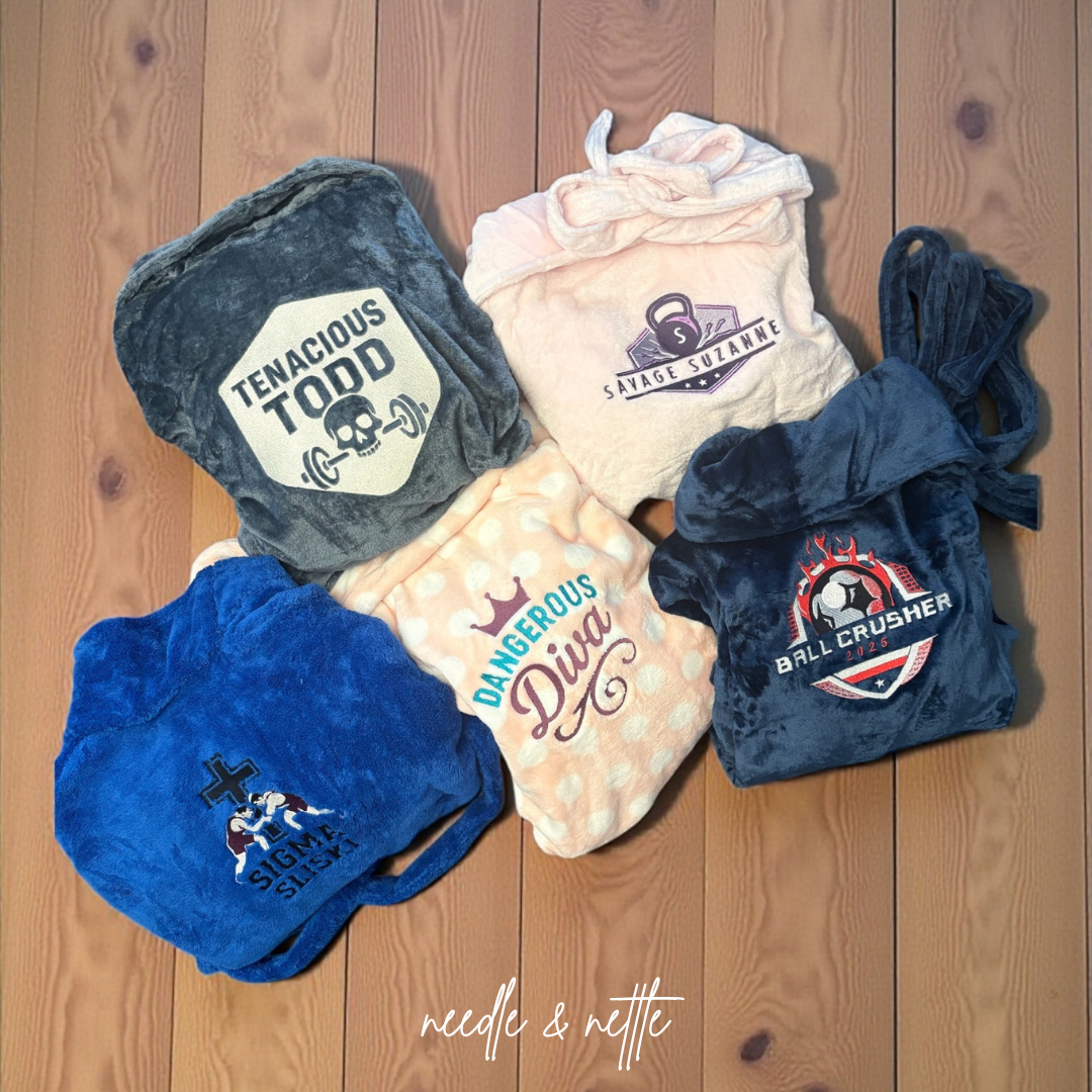 Five folded personalized towels with different designs and phrases laying on a wooden floor.