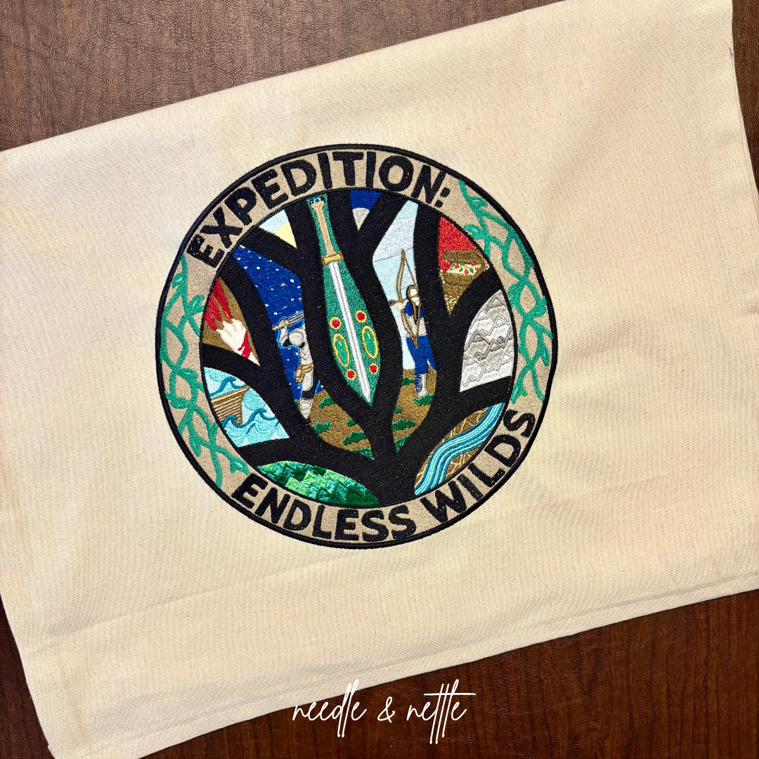 Colorful embroidered patch with the words 'Expedition: Endless Wilds' surrounding a tree with various detailed images in its branches, placed on a beige fabric background.
