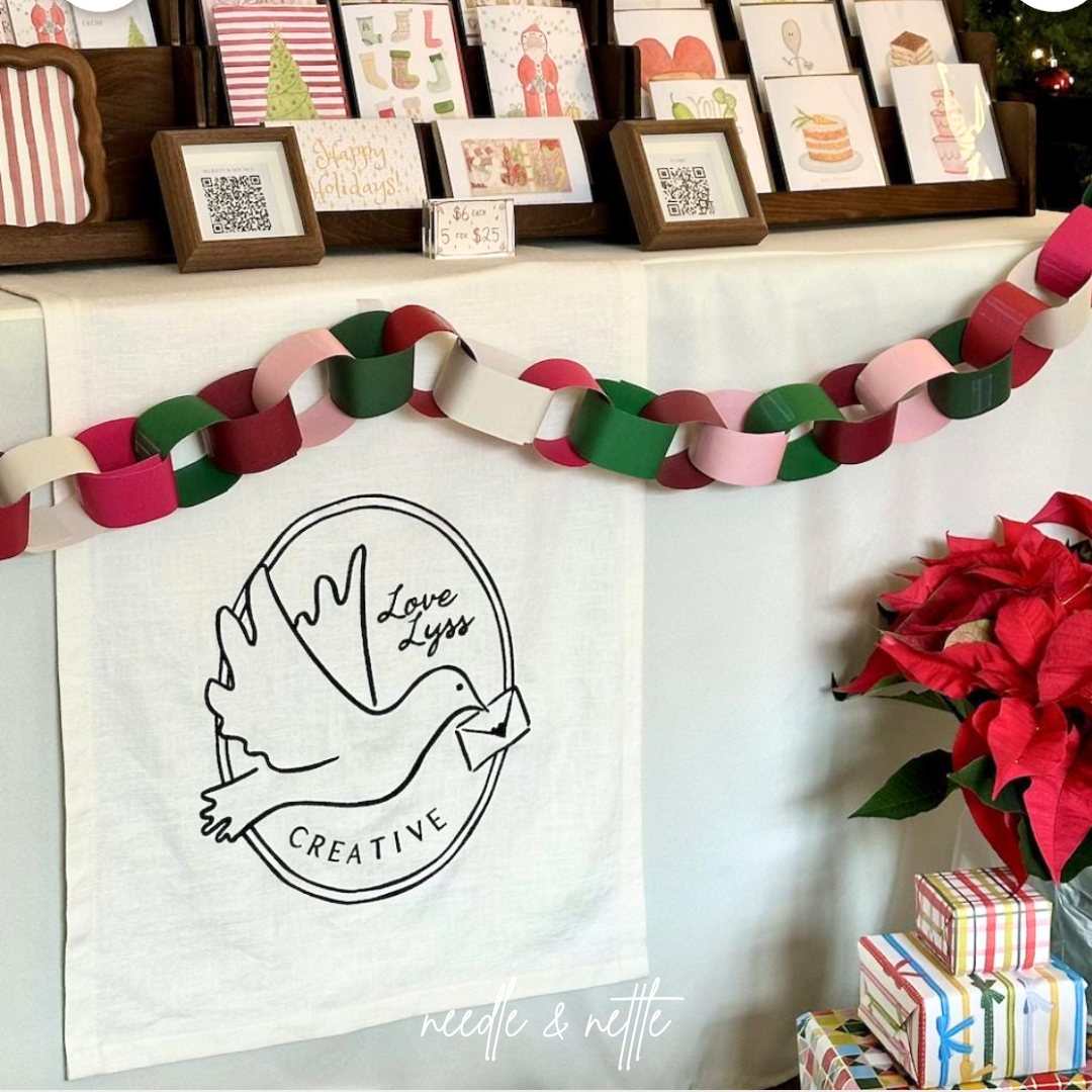 Holiday craft booth display with greeting cards, paper garland in red, pink, green, and white, poinsettia plants, and wrapped gifts, featuring a logo with a dove holding an envelope and the text "Love Lyss Creative".