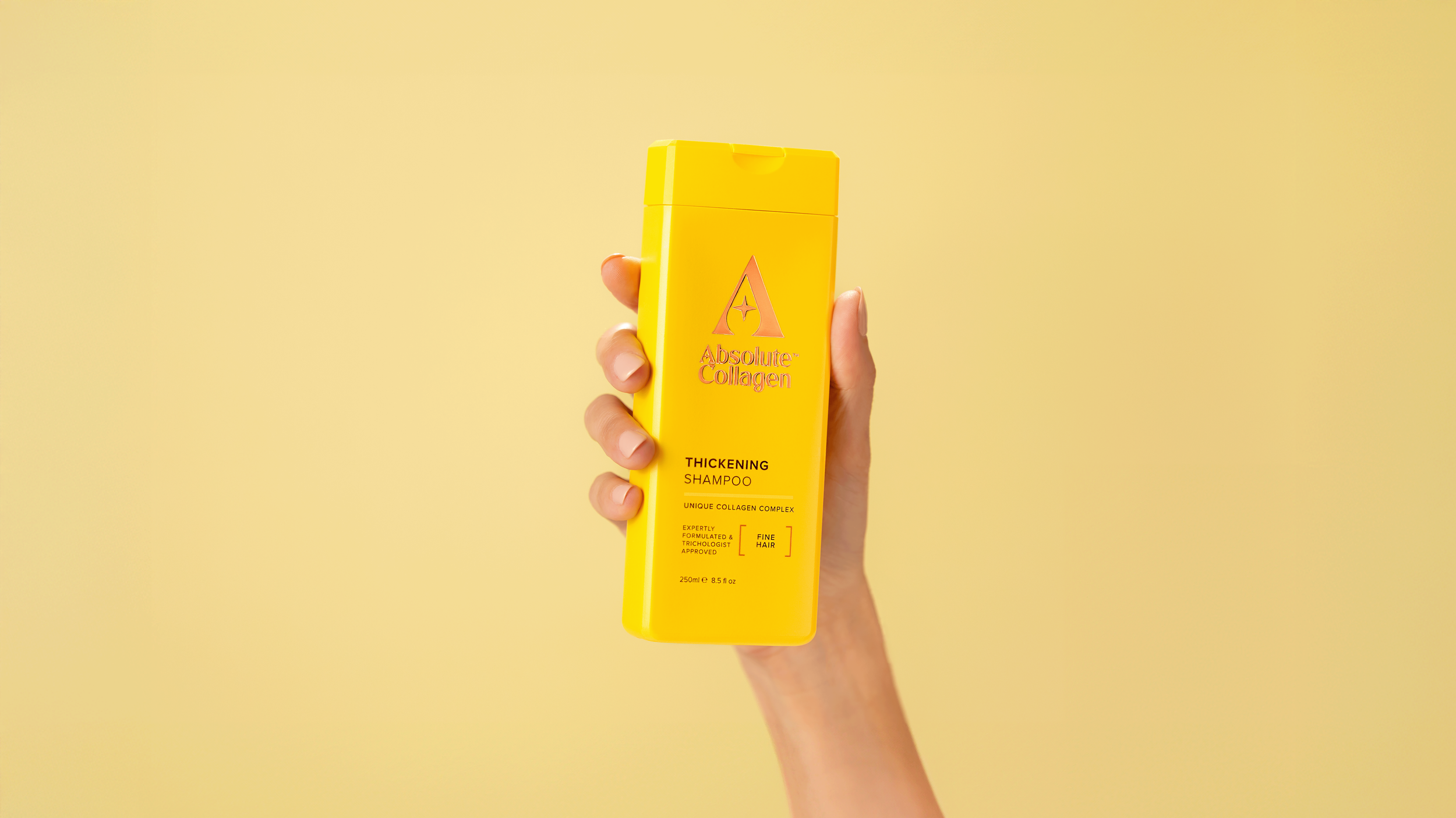 Yellow bottle of Absolute Collagen Thickening Shampoo held in a person's hand against a yellow background.