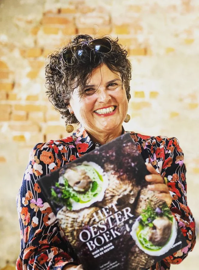 Smiling woman holding a book titled 'Het Oester Boek' with a cover showing oysters on a textured background, standing against a brick wall.